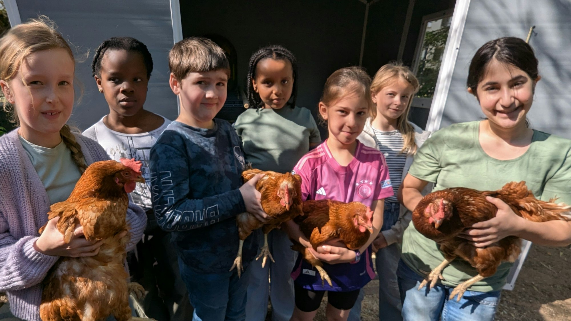 Das Foto zeigt Mädchen und Jungen vor dem Hühnerstall mit den vier Legehennen auf den Armen.