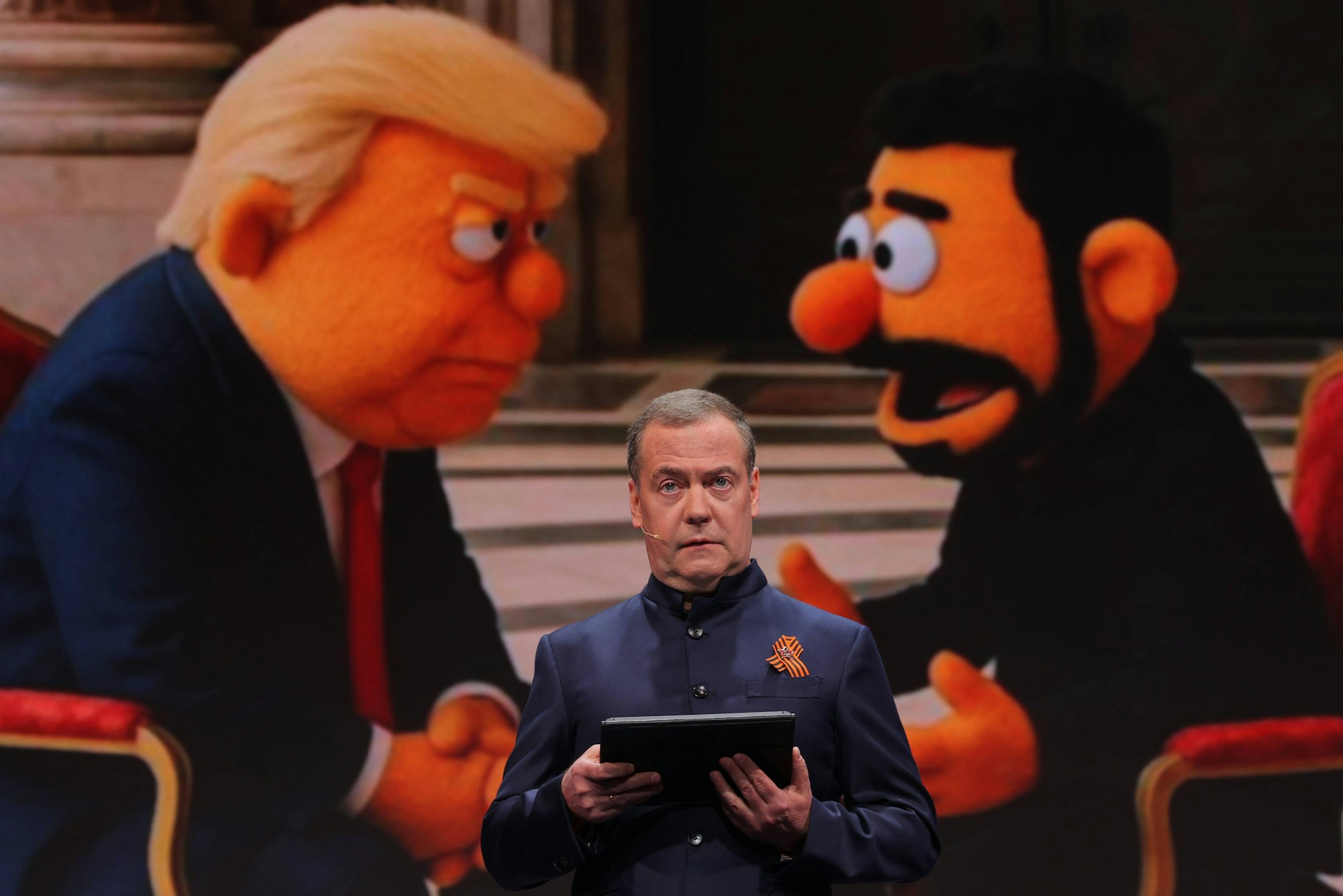 Dmitri Medwedew bei seiner Rede. Im Hintergrund sind Donald Trump und Wolodymyr Selenskyj als „Muppets“ abgebildet.