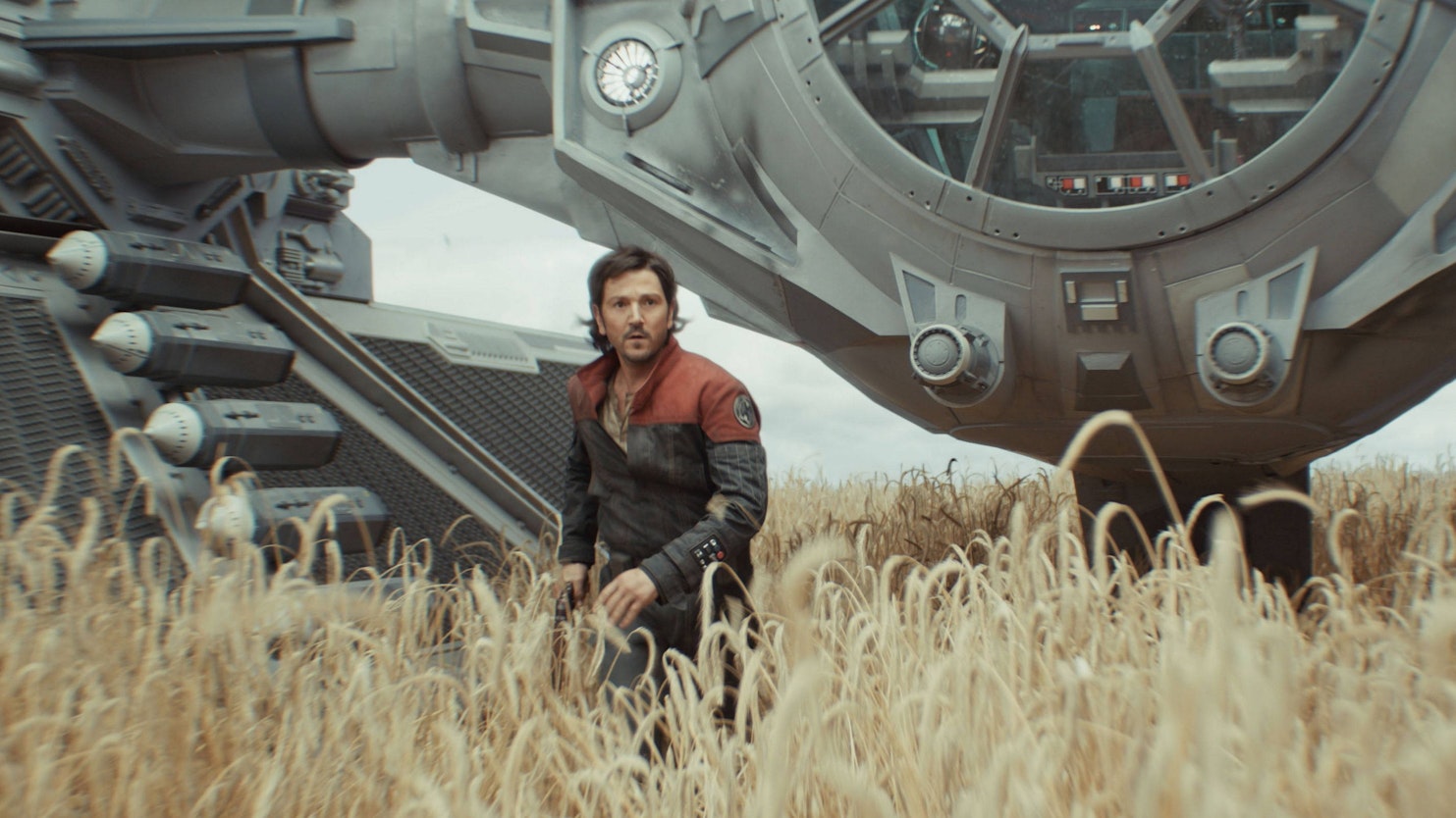 Diego Luna steht als „Cassian Andor“ in einem Weizenfeld. Hinter ihm der imperiale TIE-Fighter, den er gestohlen hat.
