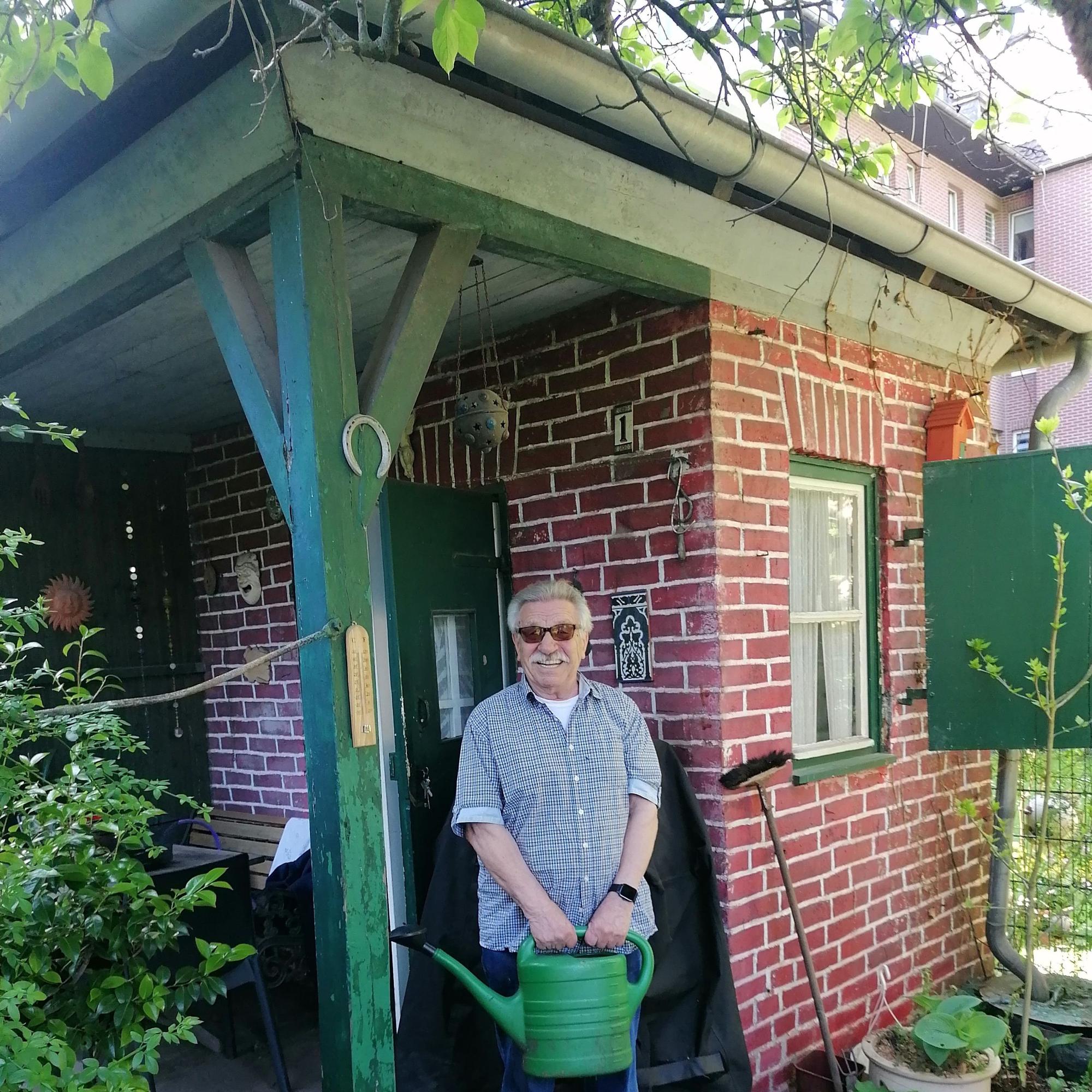 Zu sehen ist Franz Helten mit einer Gießkanne in der Hand vor seiner Gartenlaube stehen. Er lächelt in die Kamera.
