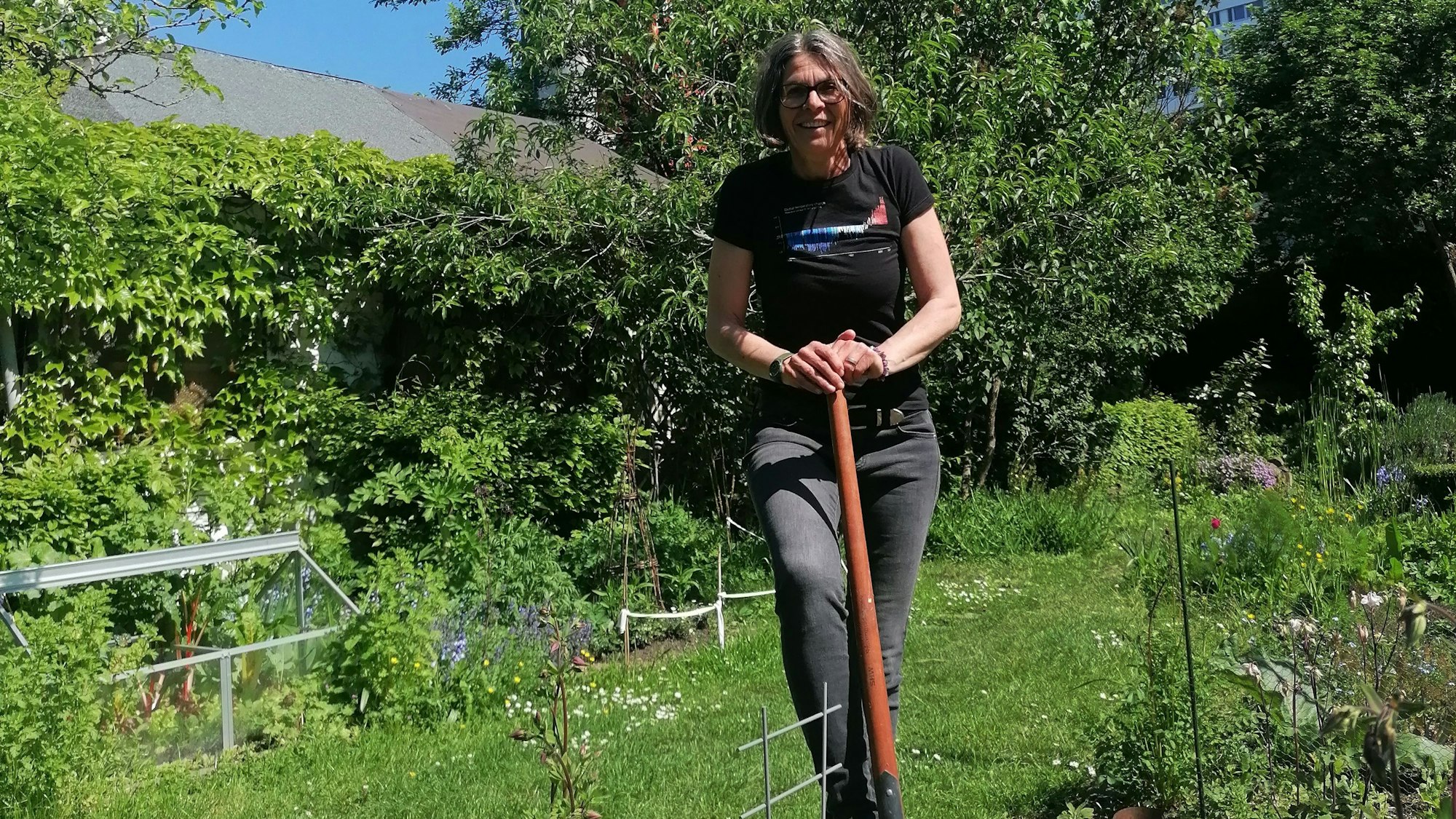 Zu sehen ist Christiane Martin in ihrem Garten in der Vorderansicht, mit einem Fuß auf einer Schaufel stehend.