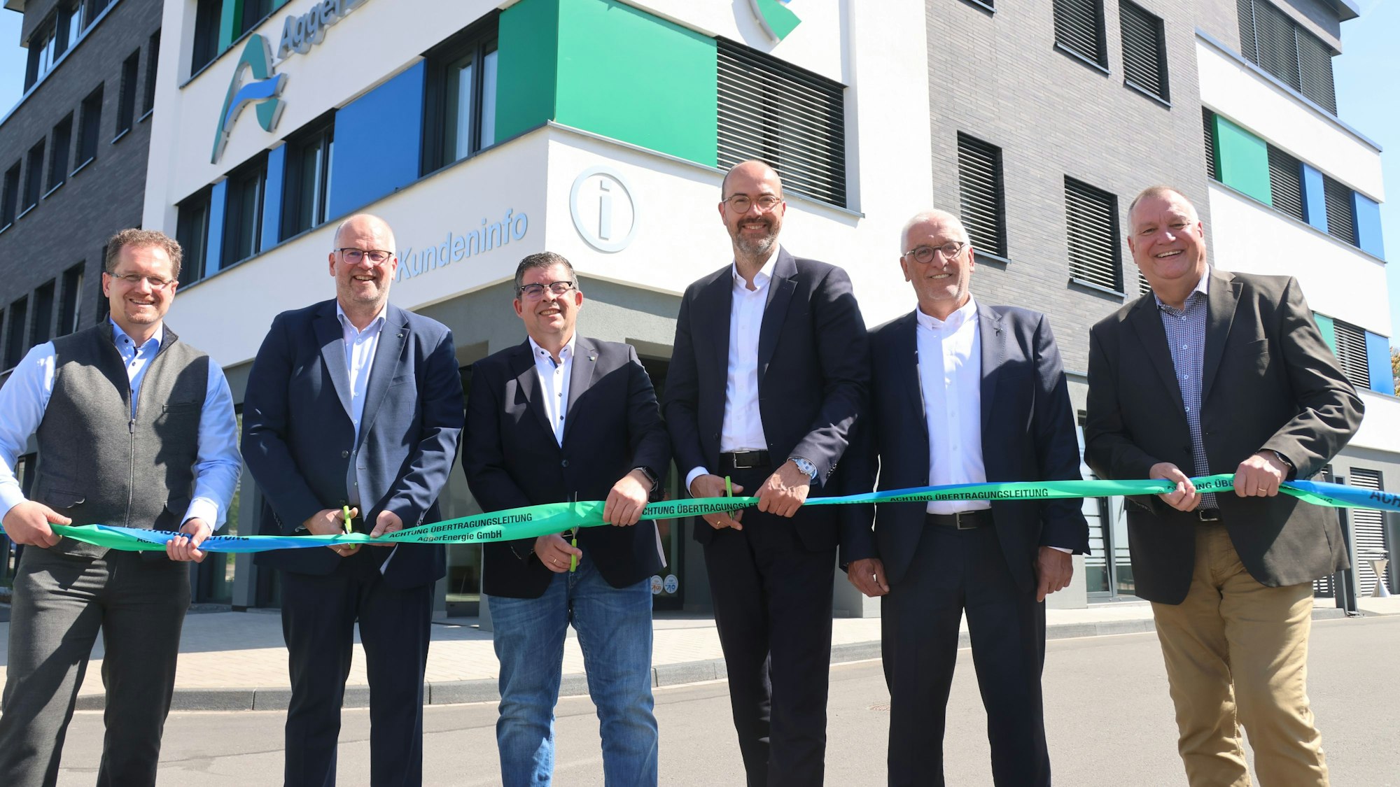 Sechs Männer schneiden ein Band vor einem neuen Gebäude der Aggerenergie durch.