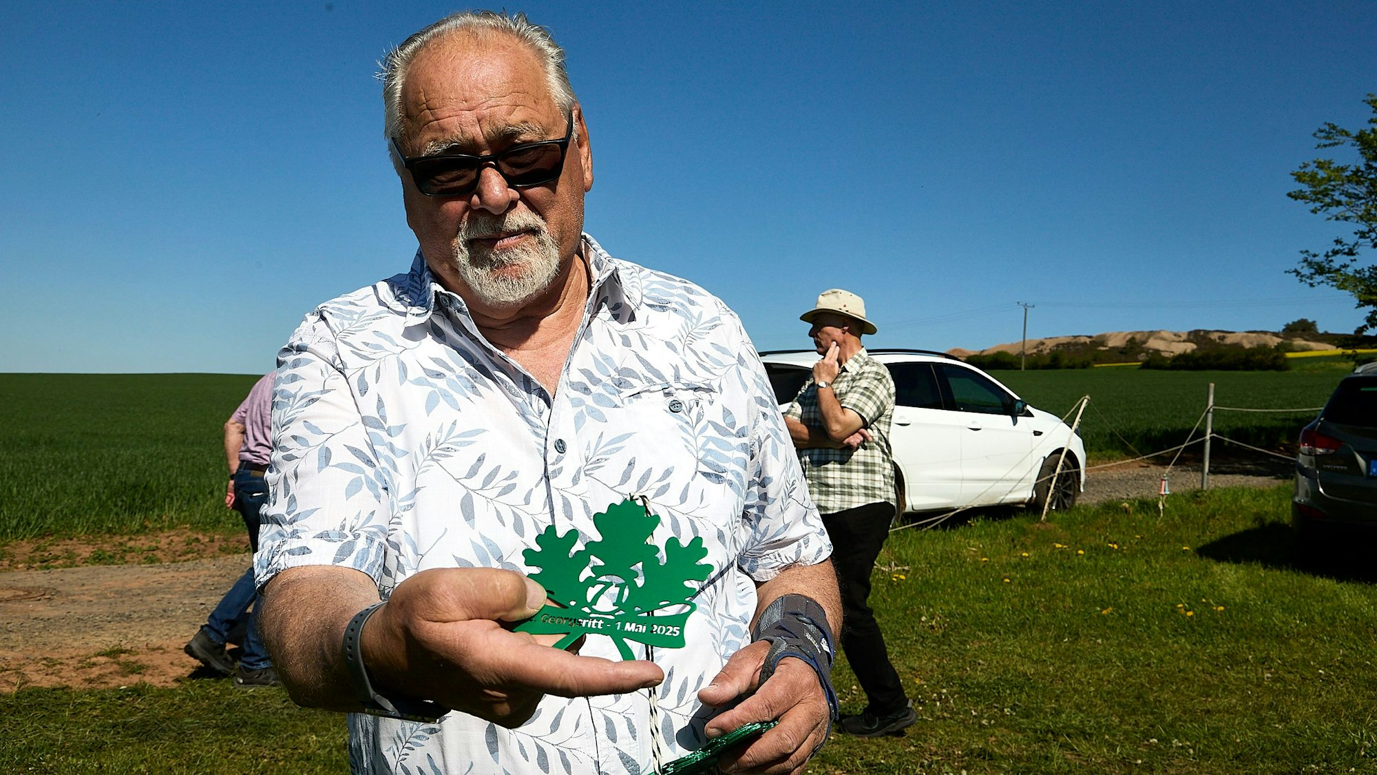 Robert Ohlerth zeigt einen grünen Orden. Er trägt ein weißes Hemd mit Blumenmuster und eine Sonnenbrille.