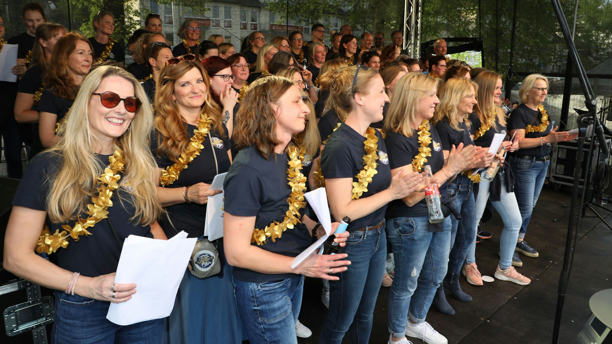 Eine Gruppe, bei denen jeder eine goldene Blumenkette trägt, steht auf einer Bühne.