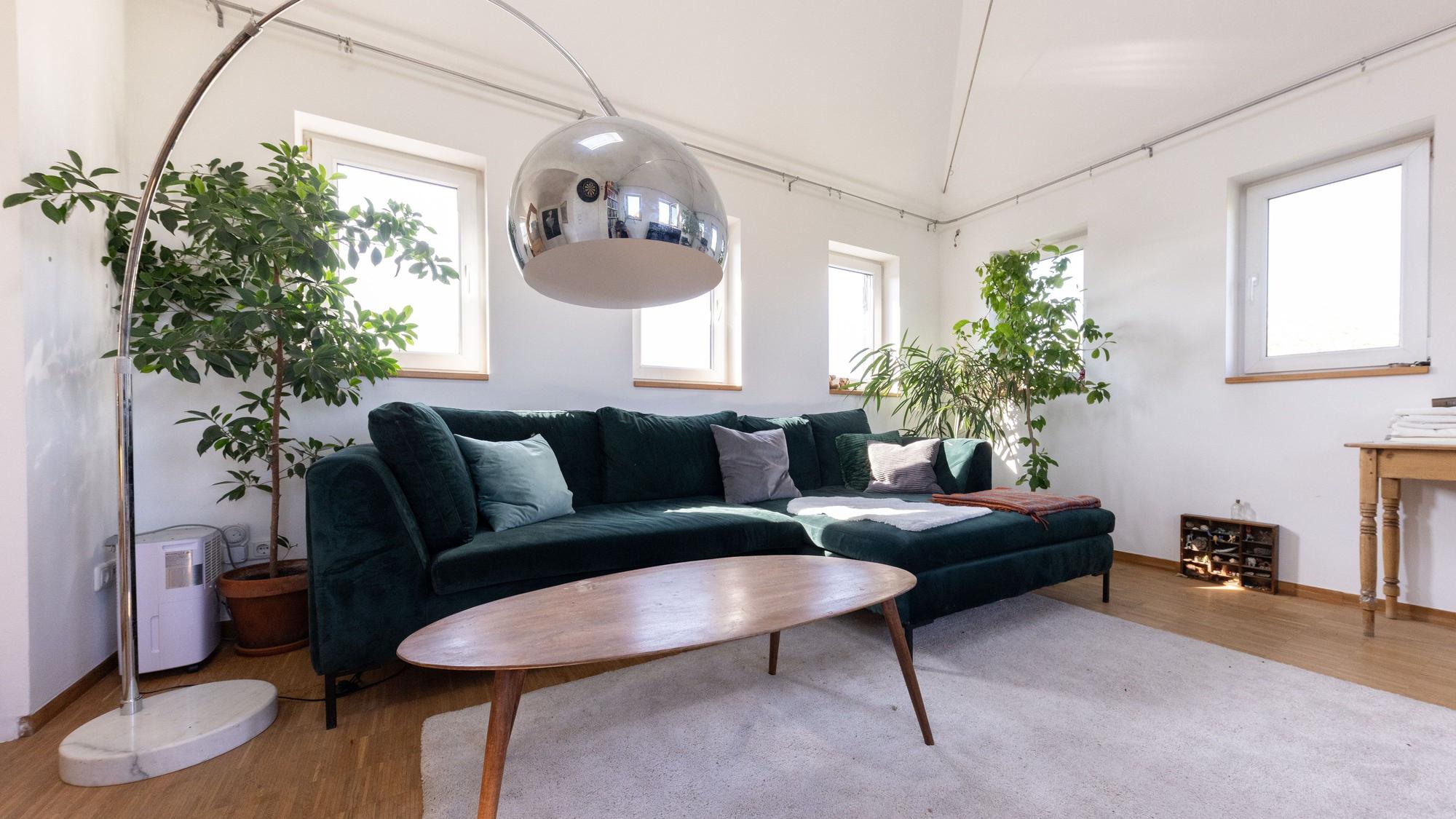 Ein Couchtisch aus Holz steht vor einem grünen Über-Eck-Sofa in einem Zimmer mit vielen Fenstern.