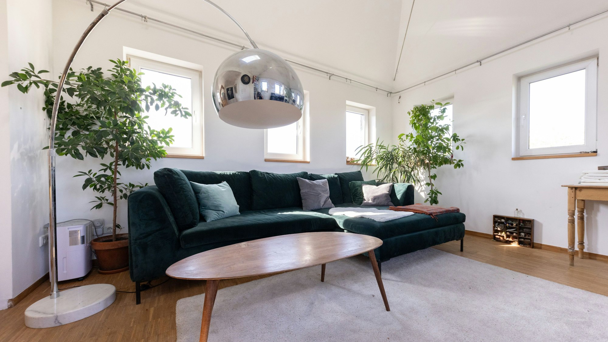 Ein Couchtisch aus Holz steht vor einem grünen Über-Eck-Sofa in einem Zimmer mit vielen Fenstern.