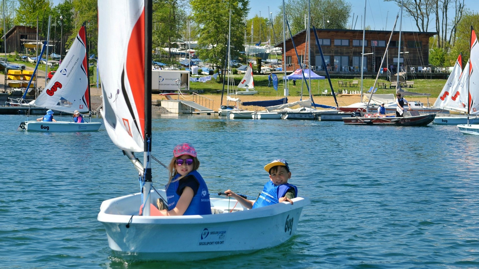 Zwei Kinder segeln über den Wassersportsee, im Hintergrund ist das Ufer zu sehen.