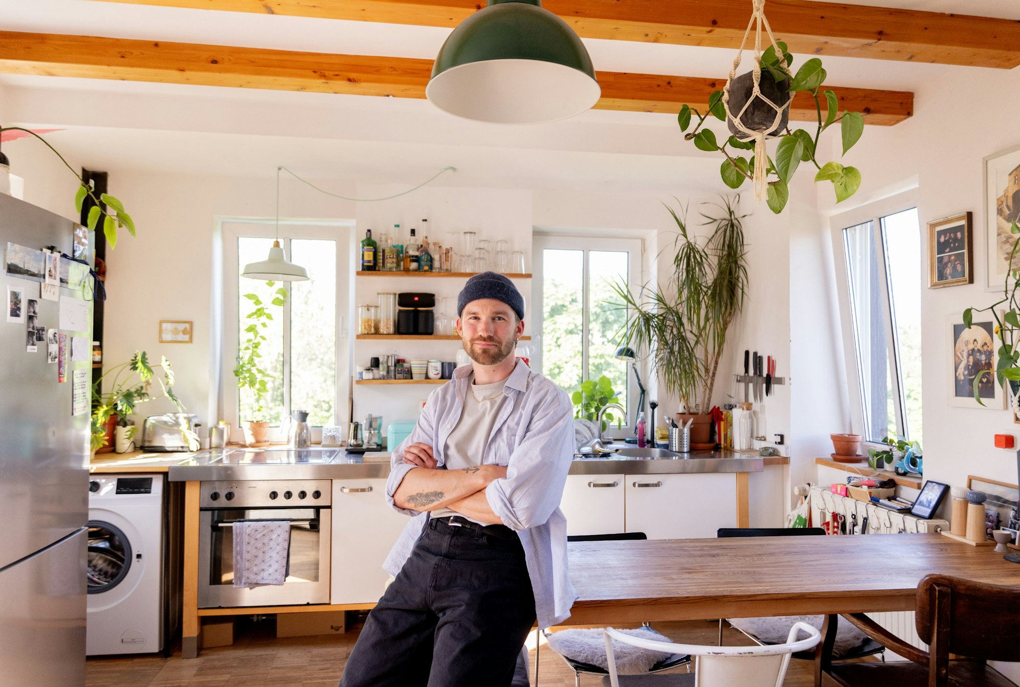 Ein junger Mann mit Strickmütze und Bart sitzt auf der Kante eines hölzernen Küchentischs.