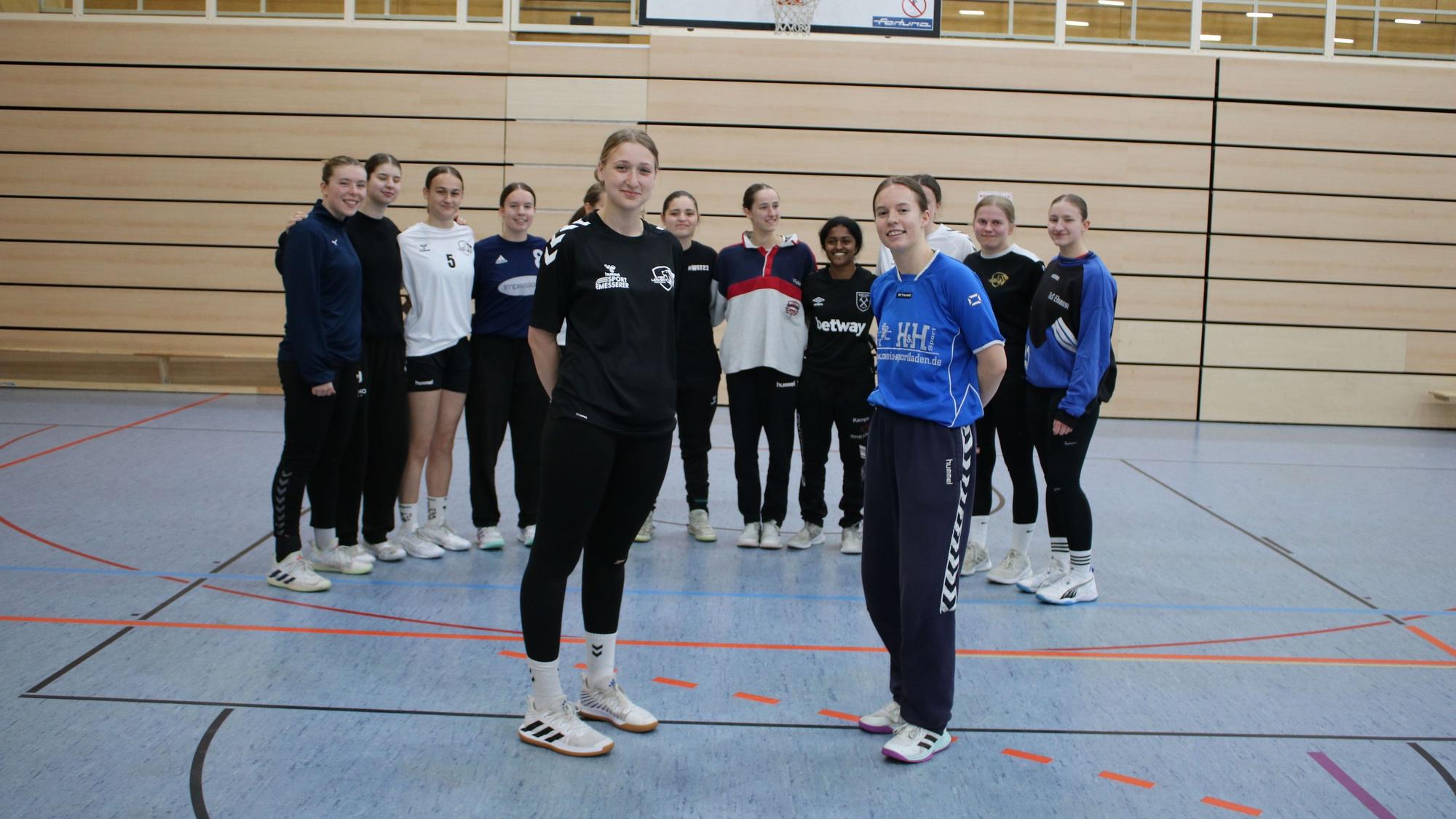 Zwei Handballerinnen stehen mit einer Mannschaft im Hintergrund in der Halle.