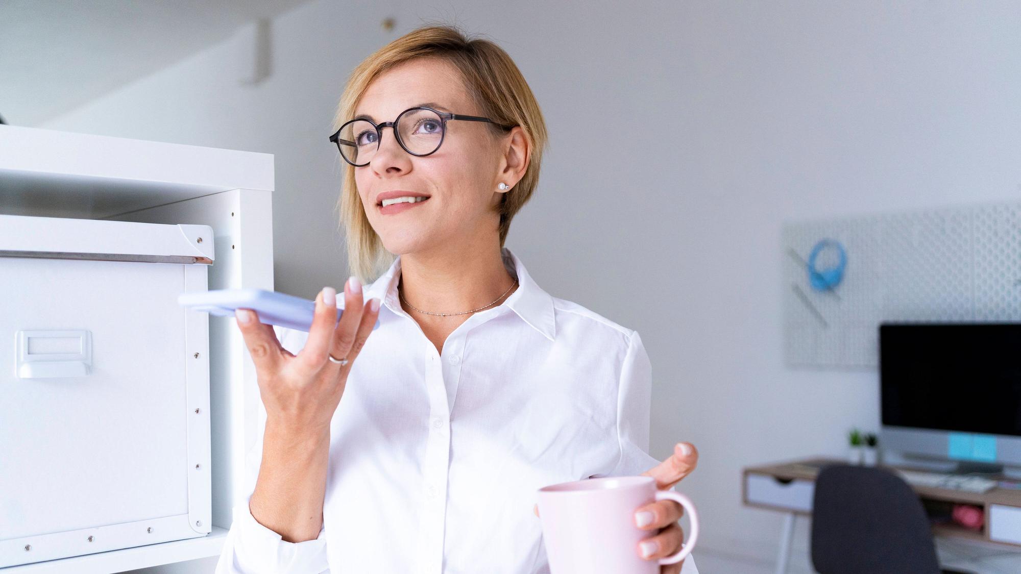Eine Frau hält in der einen Hand Kaffee und in der anderen Hand ein Handy, in das sie spricht.