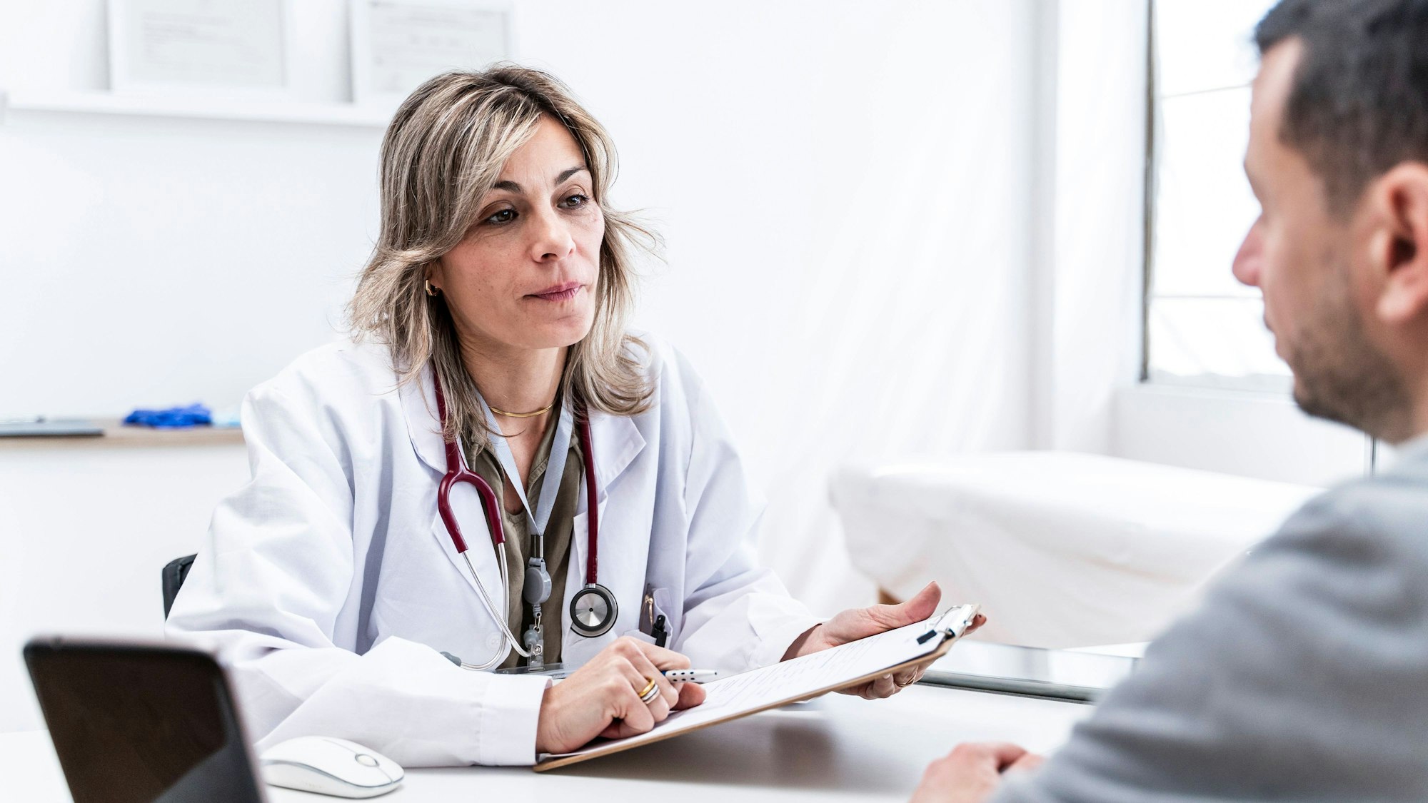 Eine Ärztin sitzt im Behandlungszimmer einer Praxis und hört ihrem Patienten zu