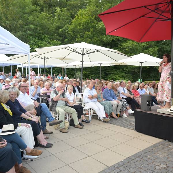 Viele Menschen sitzen unter Sonnenschirmen vor einer Bühne. Darauf einer Frau im Sommerkleid am Mikrofon.