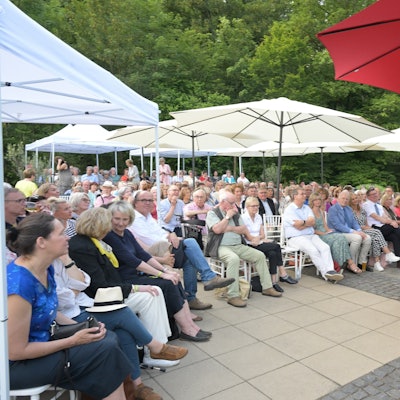 Viele Menschen sitzen unter Sonnenschirmen vor einer Bühne. Darauf einer Frau im Sommerkleid am Mikrofon.