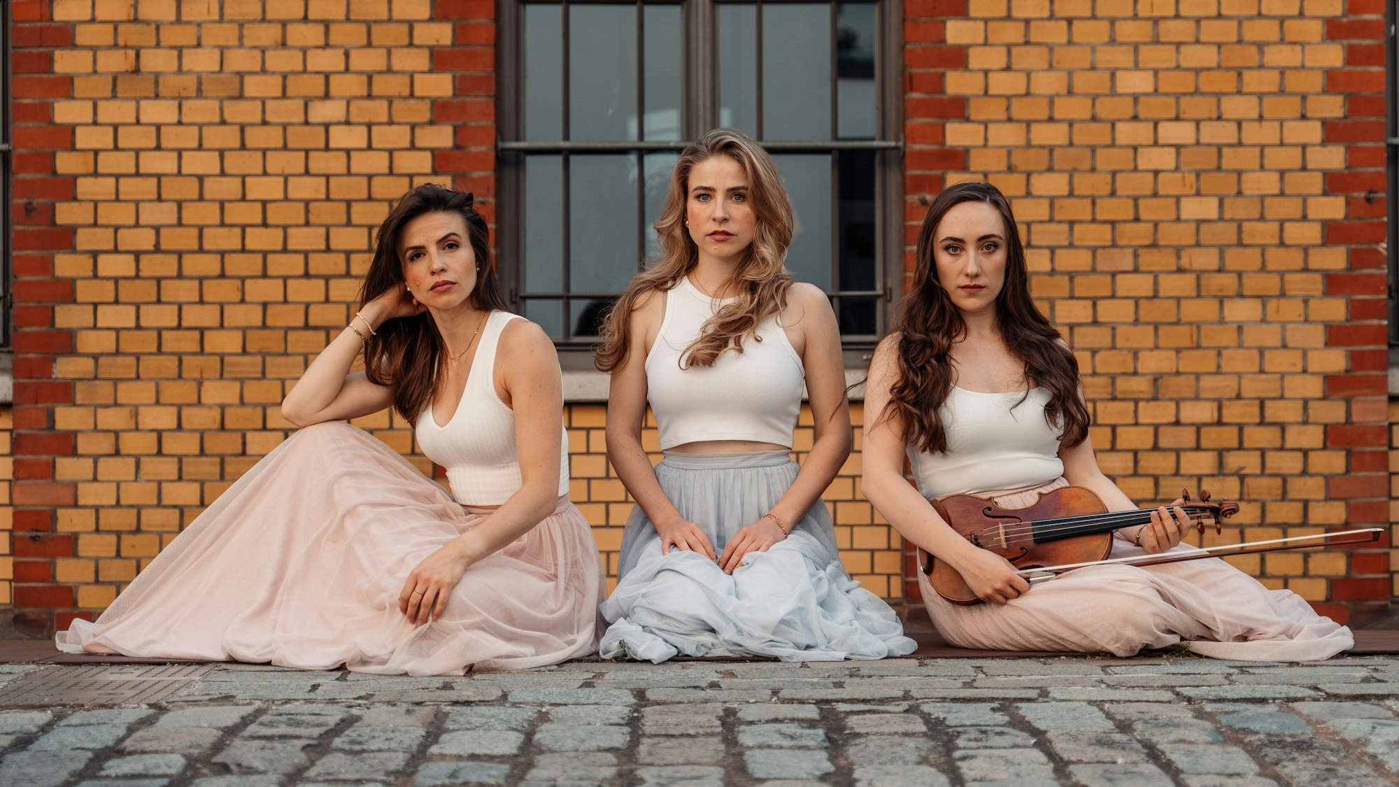 Drei Frauen, auf dem Boden sitzend. Die, die rechts sitzt, hält eine Geige und einen Geigenbogen in der Hand.