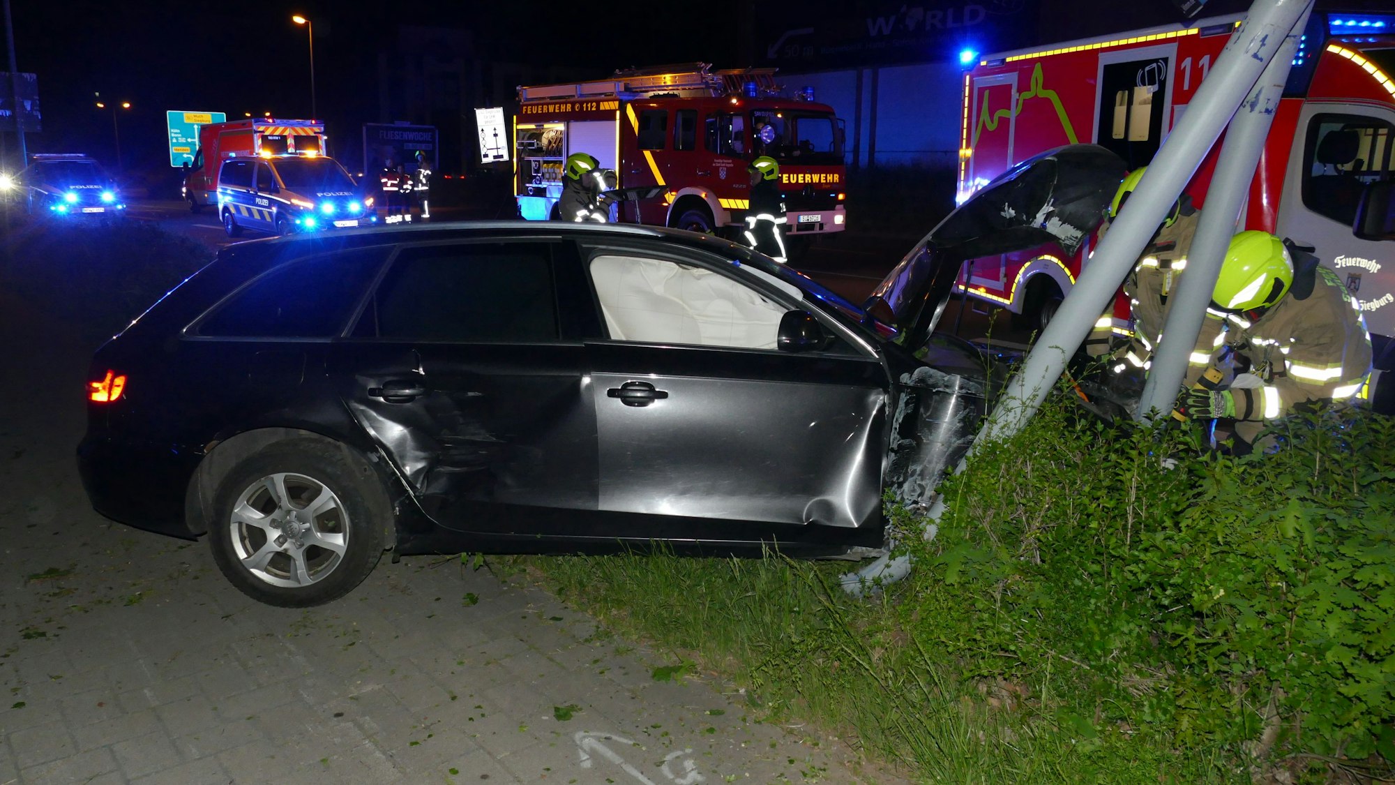 Ein dunkles Auto steht an einem Mast. Die Motorhaube ist zerstört. Im Hintergrund sind Feuerwagen zu sehen.