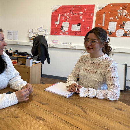 Zwei Frauen sitzen an einem Tisch und reden miteinander. Im Hintergrund hängen Plakate an den Wänden.