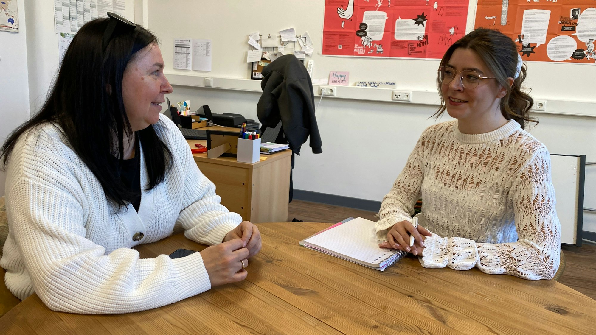 Zwei Frauen sitzen an einem Tisch und reden miteinander. Im Hintergrund hängen Plakate an den Wänden.