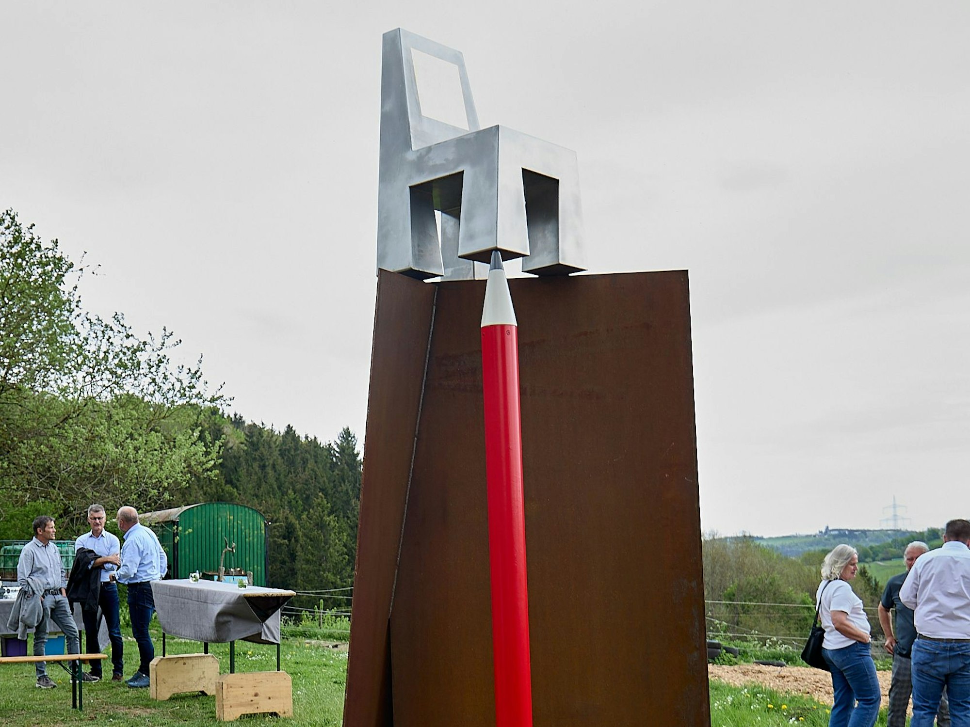 Das Kunstwerk besteht aus aufrechten Cortenstahlplatten, auf denen oben ein silberner Stuhl steht, dessen eines Bein von einem roten Bleistift gehalten wird.