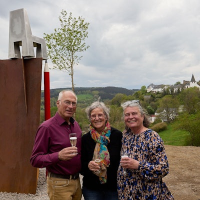 Ein Mann und zwei Frauen halten jeder ein Glas Sekt in der Hand. Links von ihnen ist eine neue Skulptur zu sehen, rechts der Blick auf den Ort Kronenburg.