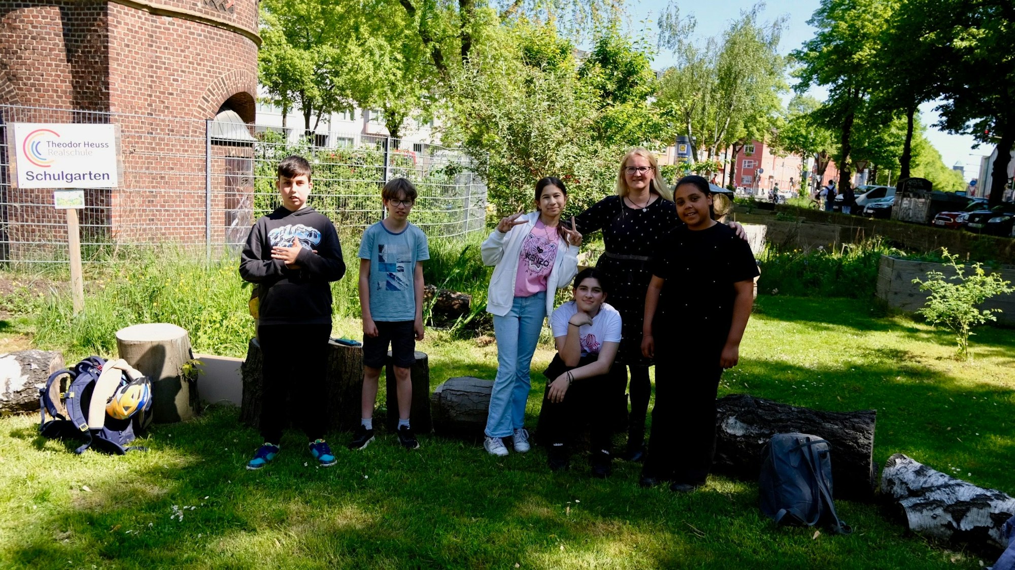 Zwei Jungs, drei Mädchen und eine Frau mit langem blondem Haar stehen in einem Garten.