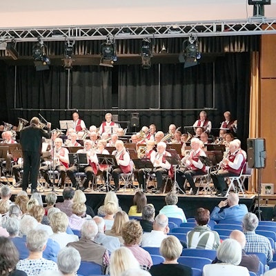 In der Aula sitzen viele Gäste, auf der Bühne sitzt das Ü-50 Blasorchester mit seinen 60 Mitgliedern.