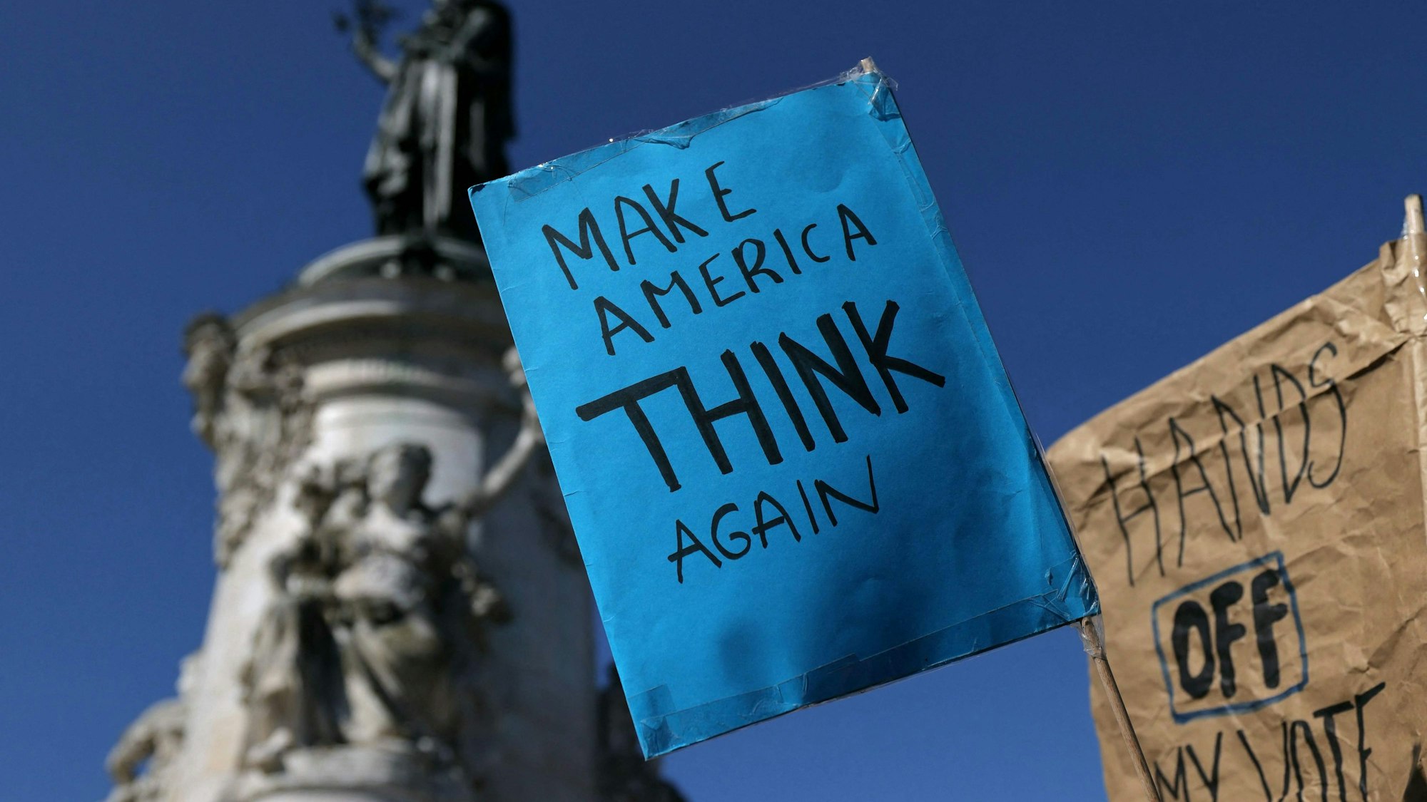 Während einer Demonstration in Paris ist ein Schild mit der Aufschrift „Make America think again“ zu sehen.