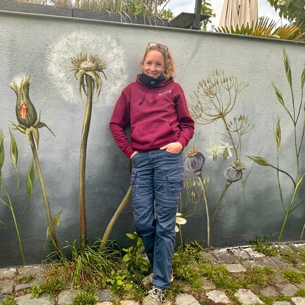 Eine Frau mit Pferdeschwanz steht vor einer Wand, die sie mit Wildblumen bemalt hat.