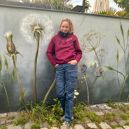 Eine Frau mit Pferdeschwanz steht vor einer Wand, die sie mit Wildblumen bemalt hat.