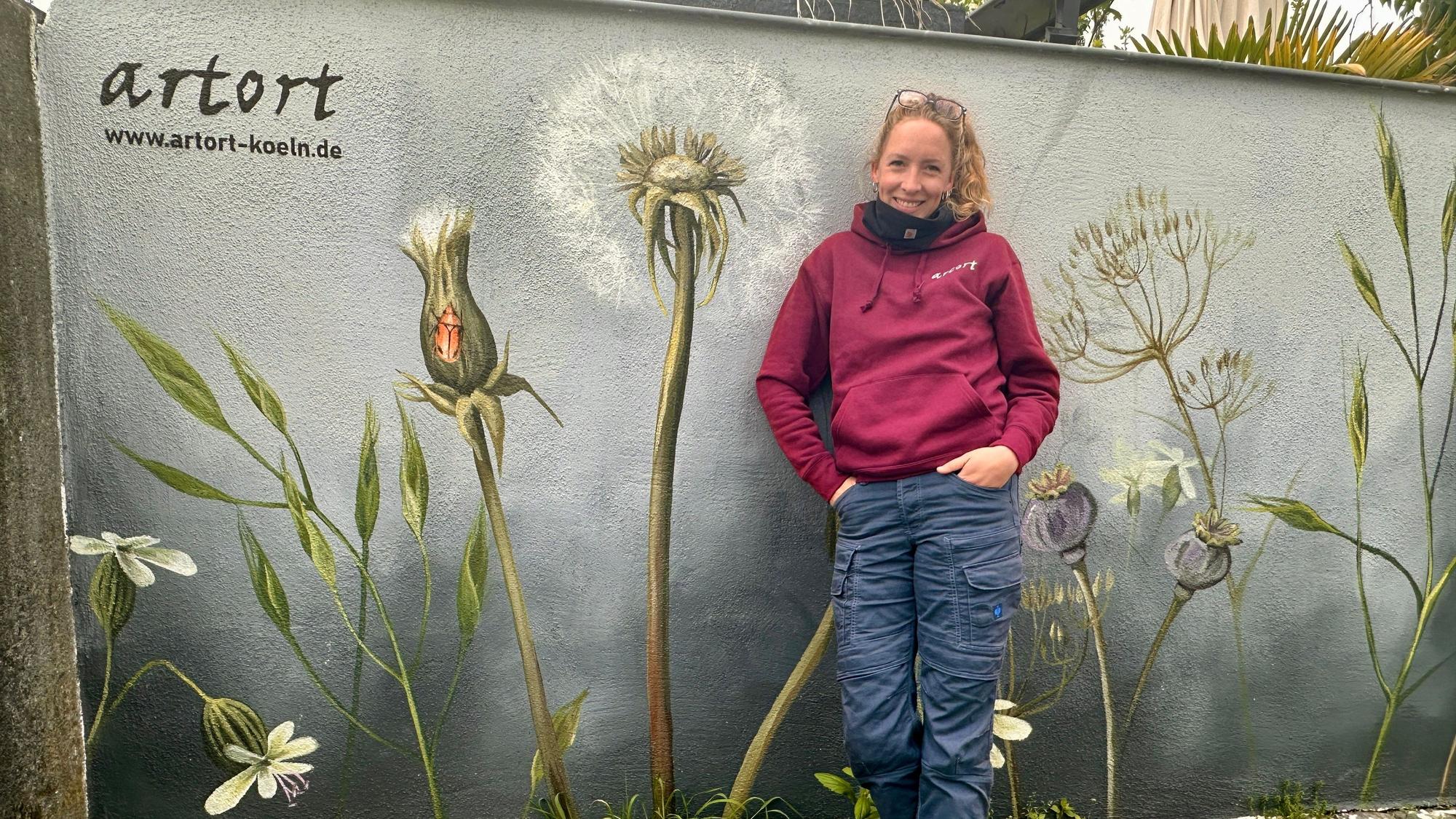 Eine Frau mit Pferdeschwanz steht vor einer Wand, die sie mit Wildblumen bemalt hat.