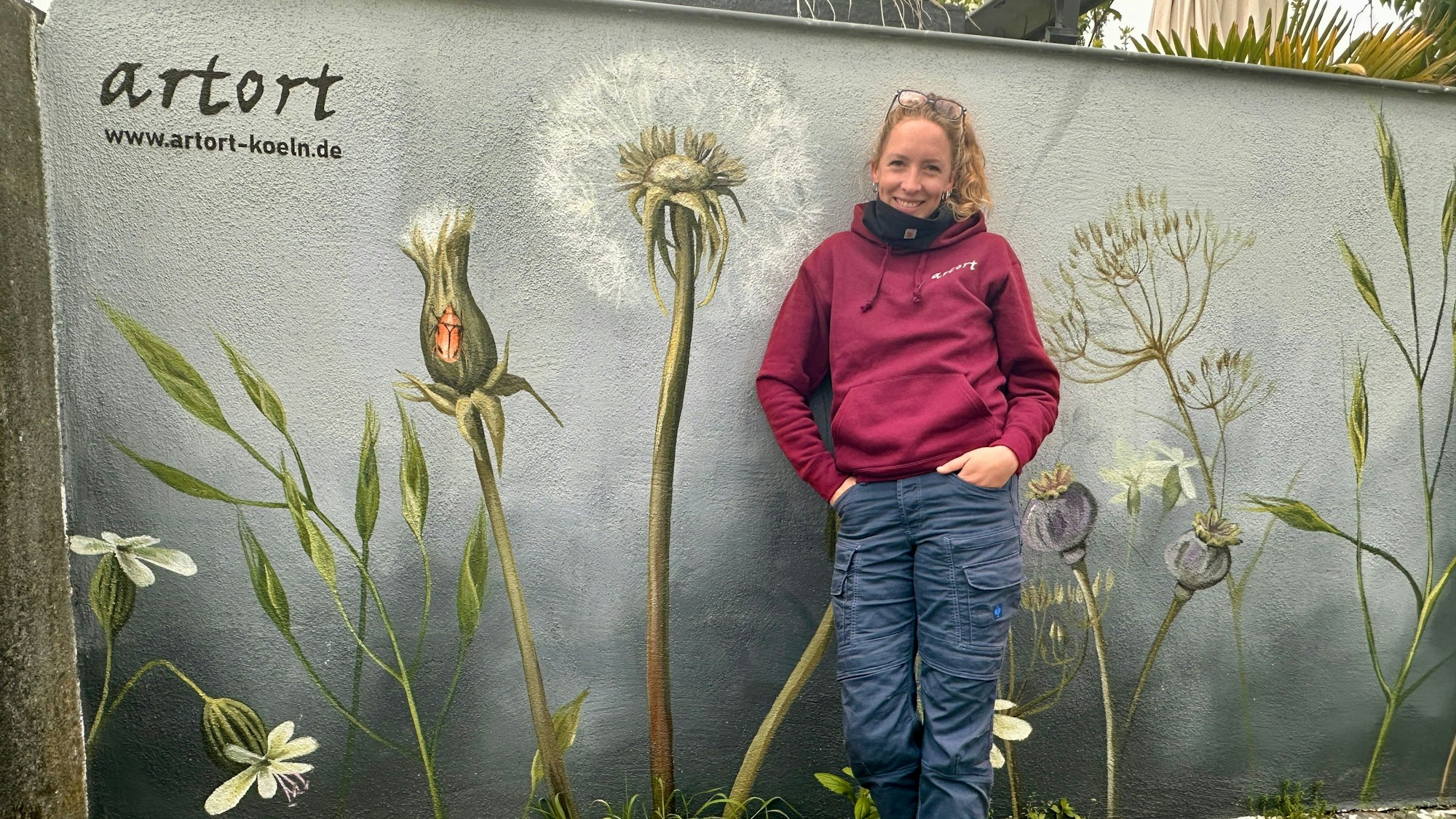 Eine Frau mit Pferdeschwanz steht vor einer Wand, die sie mit Wildblumen bemalt hat.