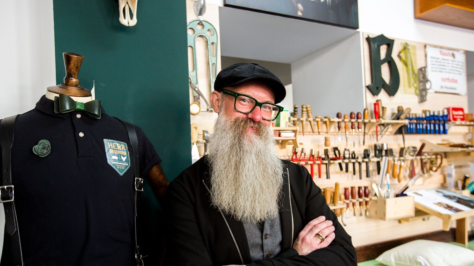 Ein Mann mit Brille und langem grauen Bart steht mit verschränkten Armen vor einer Werkbank mit allerlei Werkzeugen.