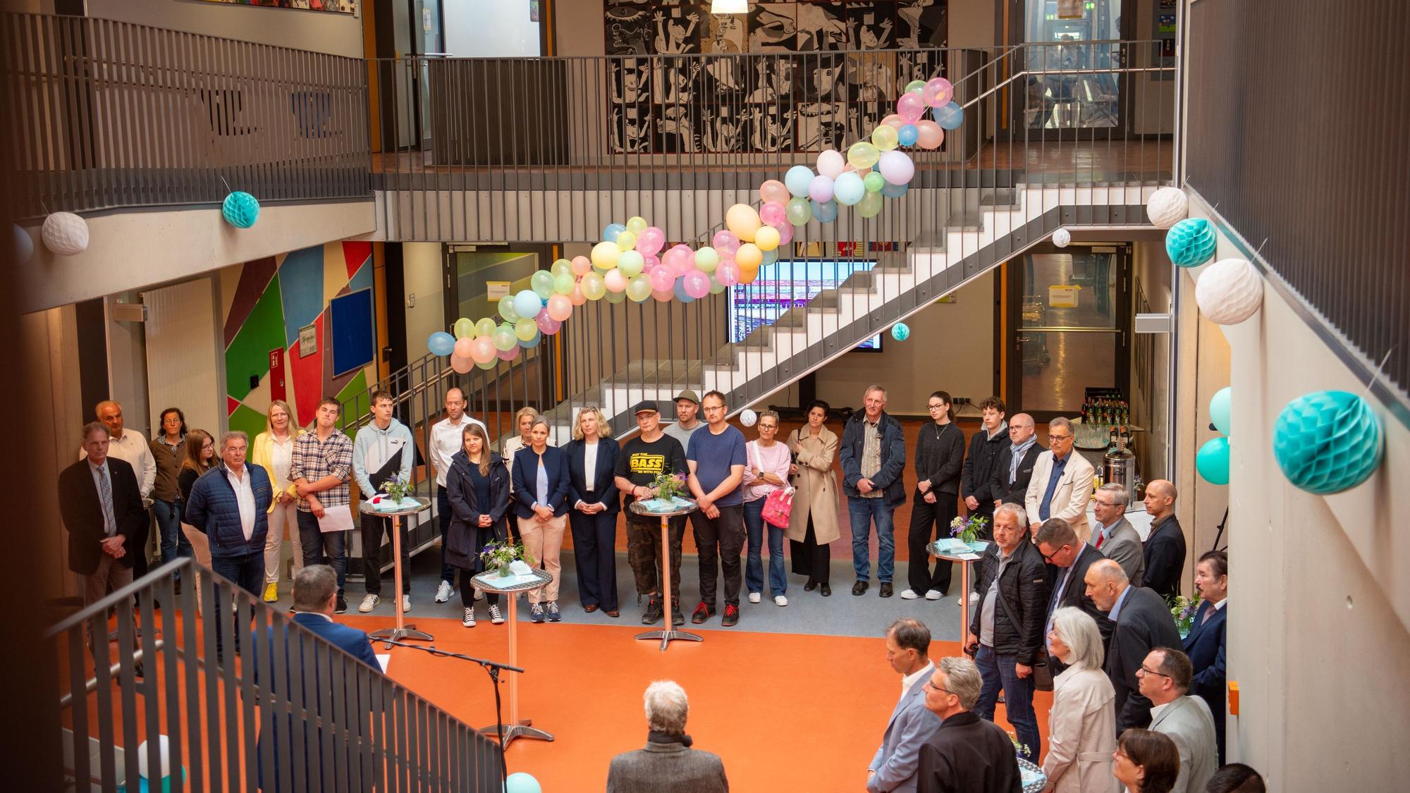 Einige Menschen stehen in einem mit Blumen und Luftballons geschmückten Foyer und hören einem Redner zu.