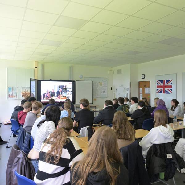 Schüler sitzen in ihrem Klassenzimmer und folgen einer Videokonferenz auf der großen Leinwand.