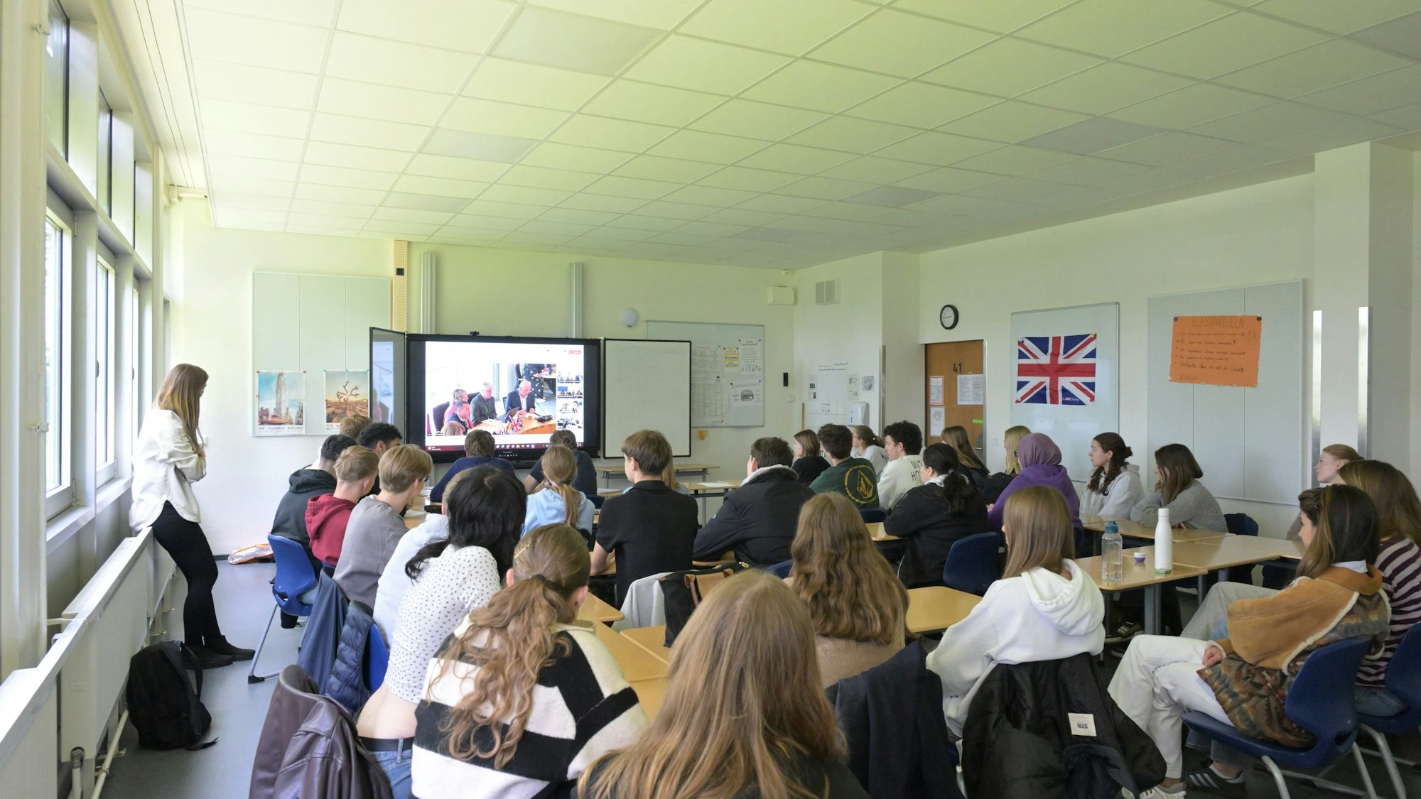 Schüler sitzen in ihrem Klassenzimmer und folgen einer Videokonferenz auf der großen Leinwand.