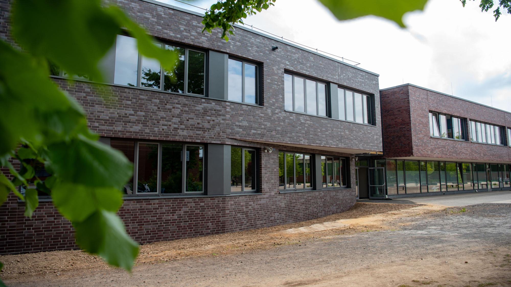 Hinter grünen Ästen sieht man ein modernes Schulgebäude mit rot-brauner Verkleidung.
