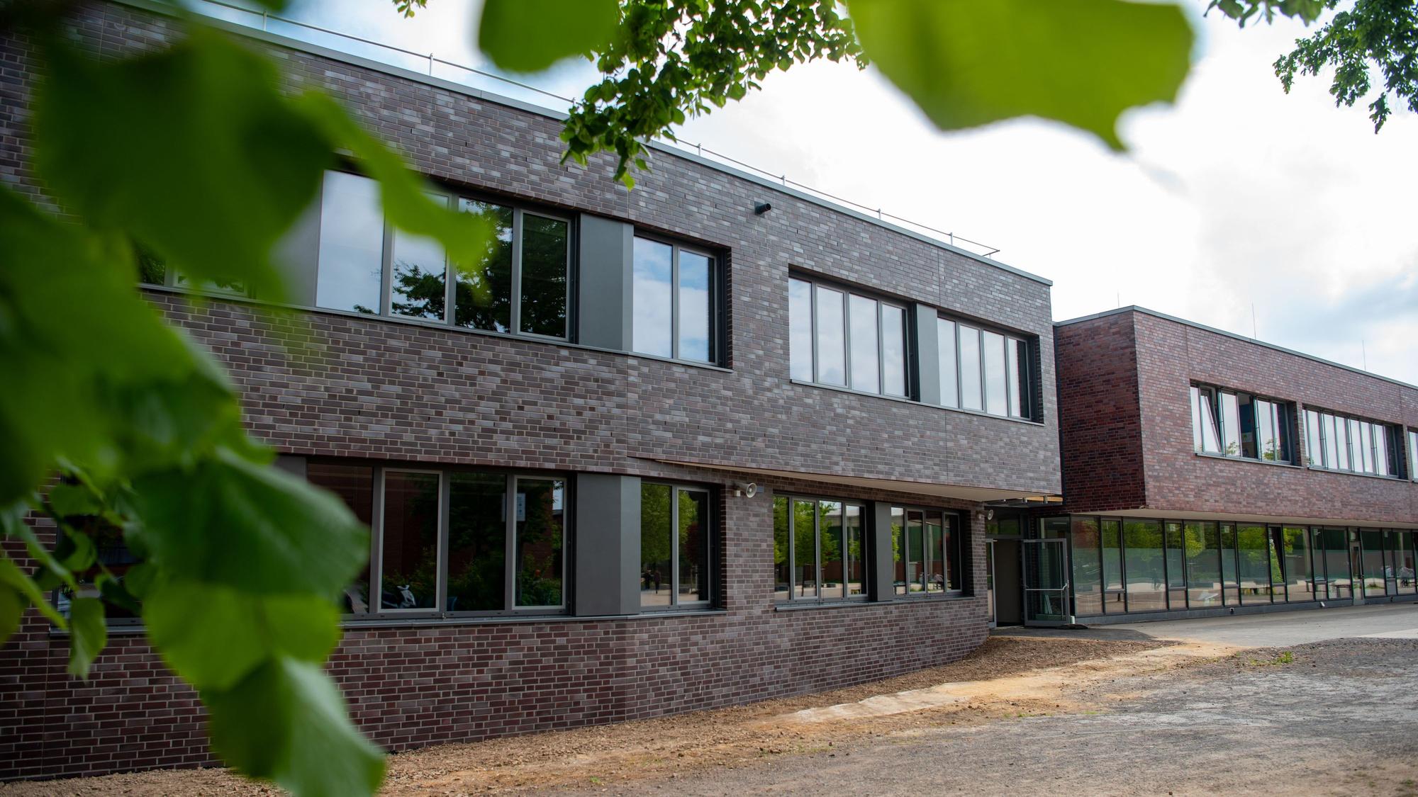 Hinter grünen Ästen sieht man ein modernes Schulgebäude mit rot-brauner Verkleidung.