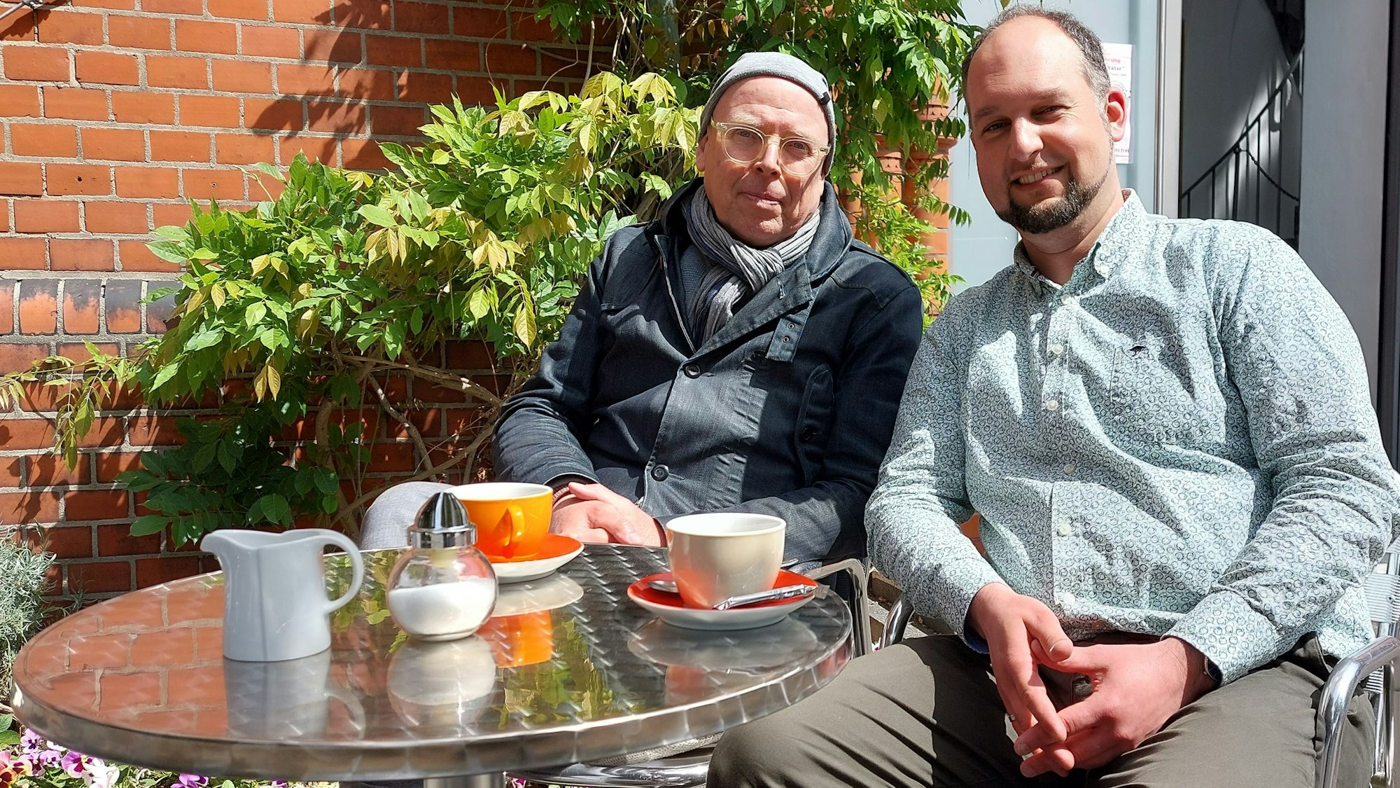 Zwei Männer sitzen an einem kleinen Bistrotisch vor einem Backsteingebäude.