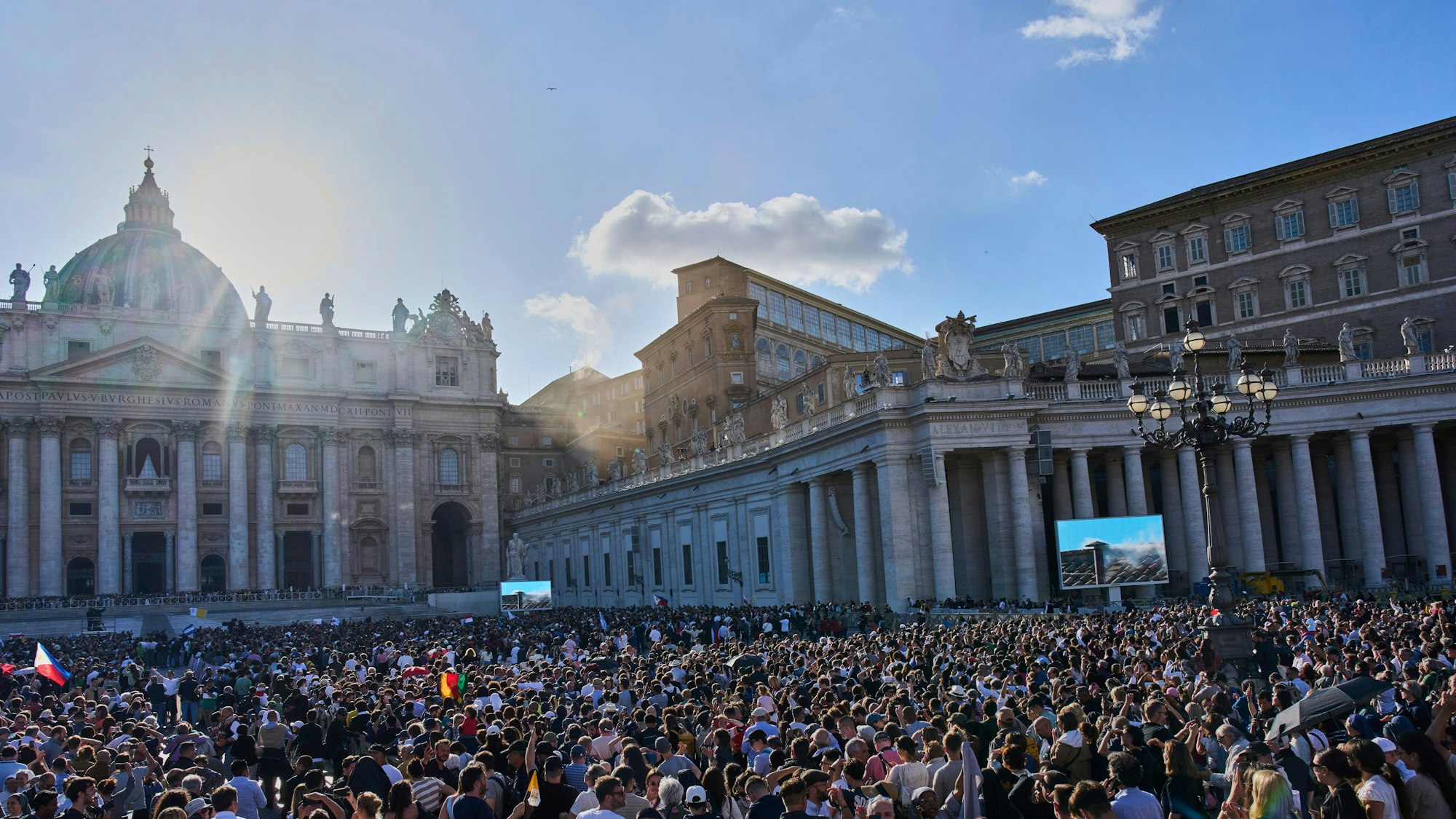 Das Foto zeigt Menschen, die auf dem Petersplatz auf einer Videoleinwand verfolgen, wie weißer Rauch aus dem Schornstein der Sixtinischen Kapelle aufsteigt. Foto: Bernat Armangue/AP/dpa