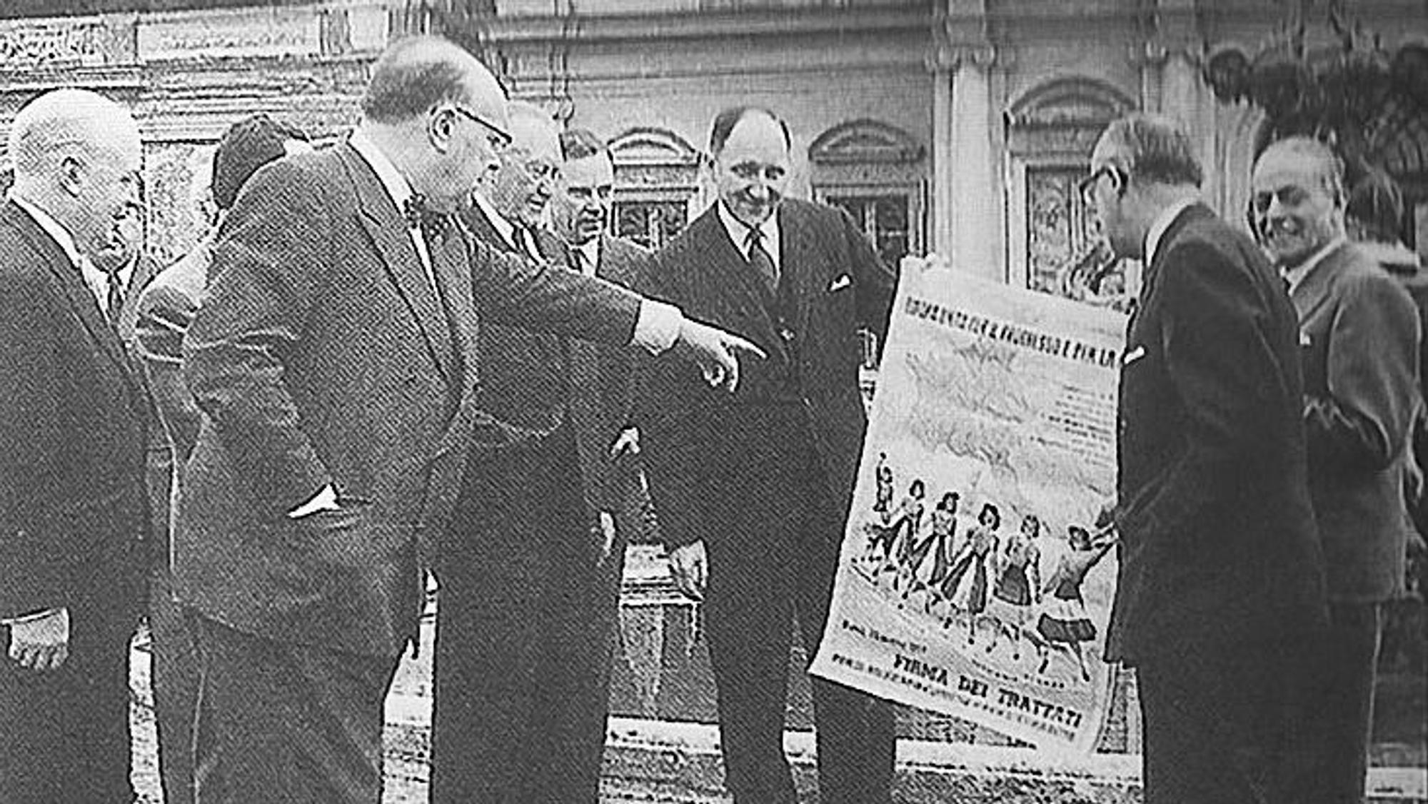 Ein altes Schwarz-Weiß-Foto zeigt Männer in Anzügen, darunter auch Bundeskanzler Konrad Adenauer, die ein Plakat betrachten.