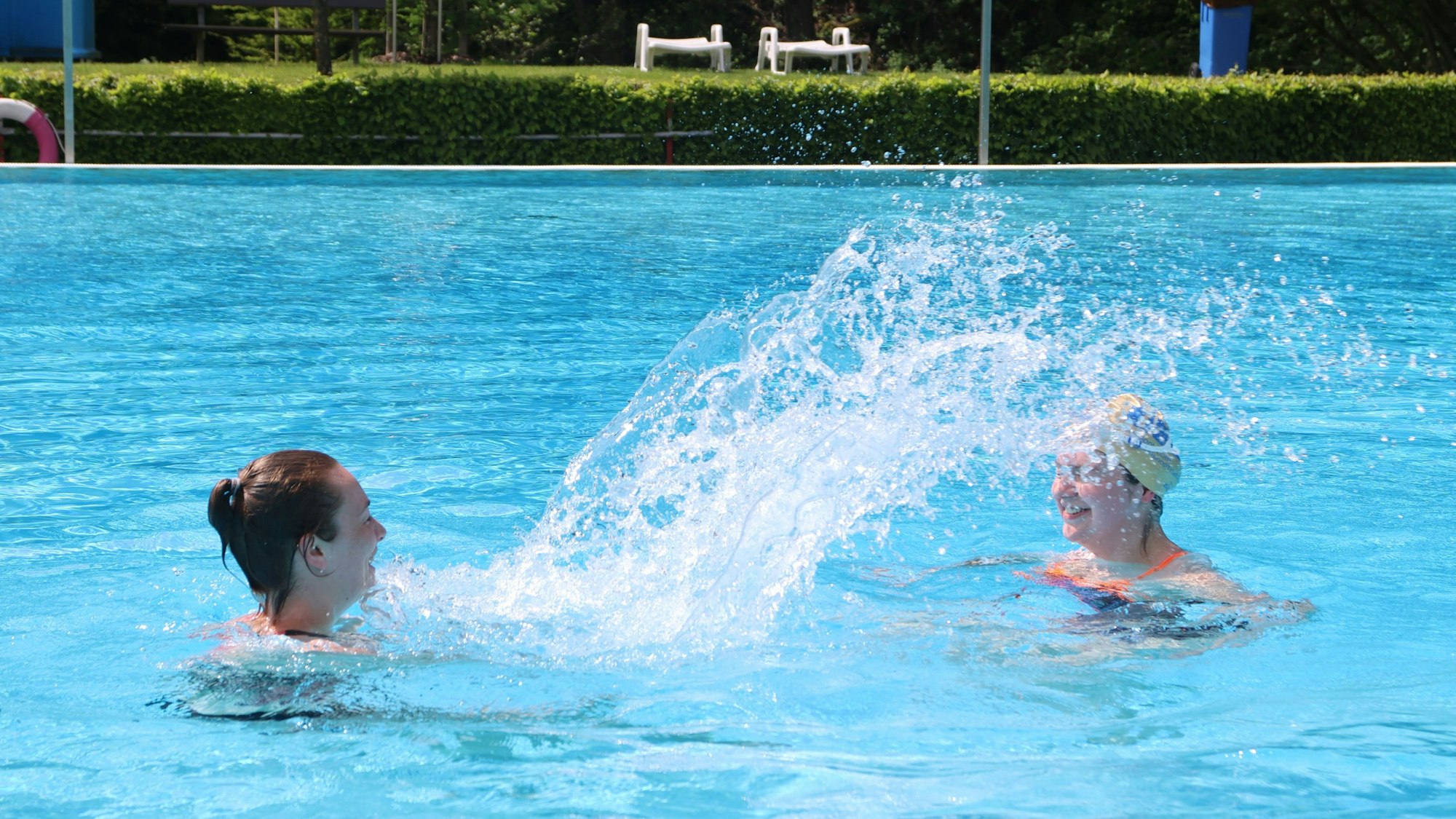 Zwei Frauen schwimmen in einem Freibad und spritzen sich Wasser entgegen.