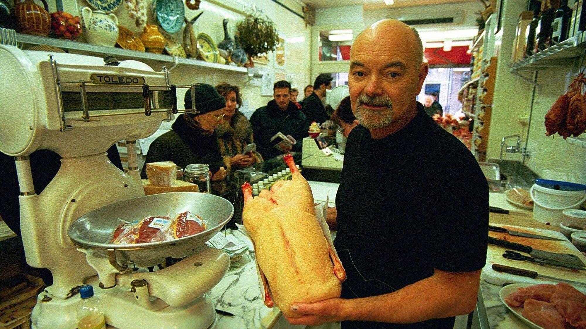 Hans Georg Rochow hält ein Geflügelprodukt in die Kamera. Im Hintergrund stehen mehre Kunden an einer Ladentheke.