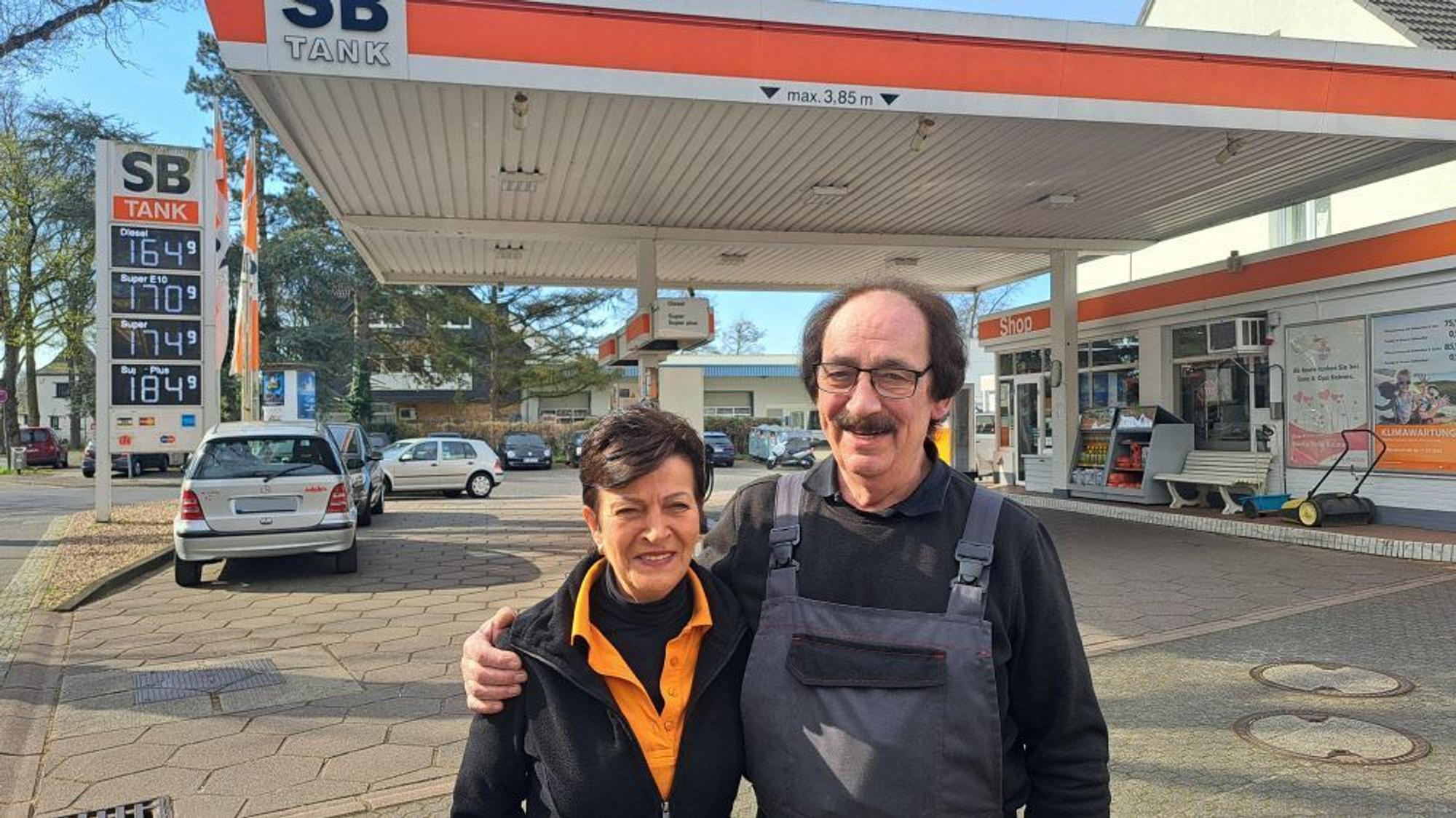 Eine Frau und ein Mann mit schwarzen Haaren stehen vor einer Tankstelle.