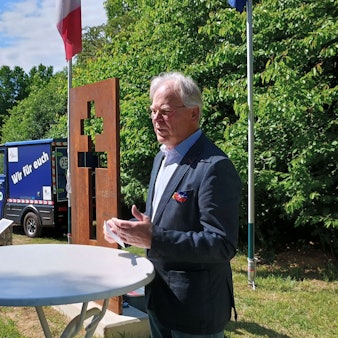 Hübner steht schräg vor dem Lothringer Kreuz. Das Kreuz hat zwei Querbalken und ist als Lücke in einer rostigen Stahlplatte sichtbar.
