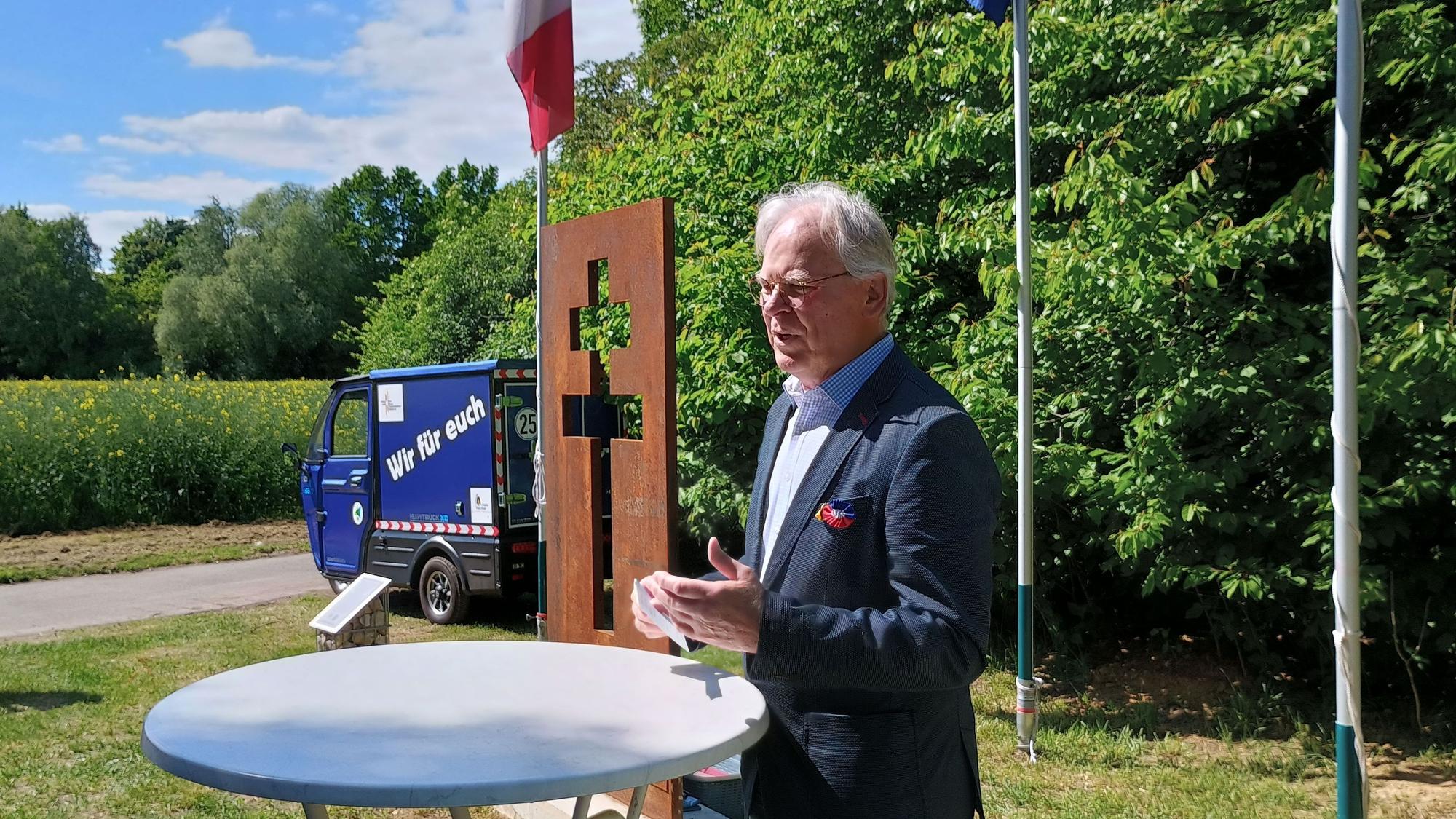 Hübner steht schräg vor dem Lothringer Kreuz. Das Kreuz hat zwei Querbalken und ist als Lücke in einer rostigen Stahlplatte sichtbar.