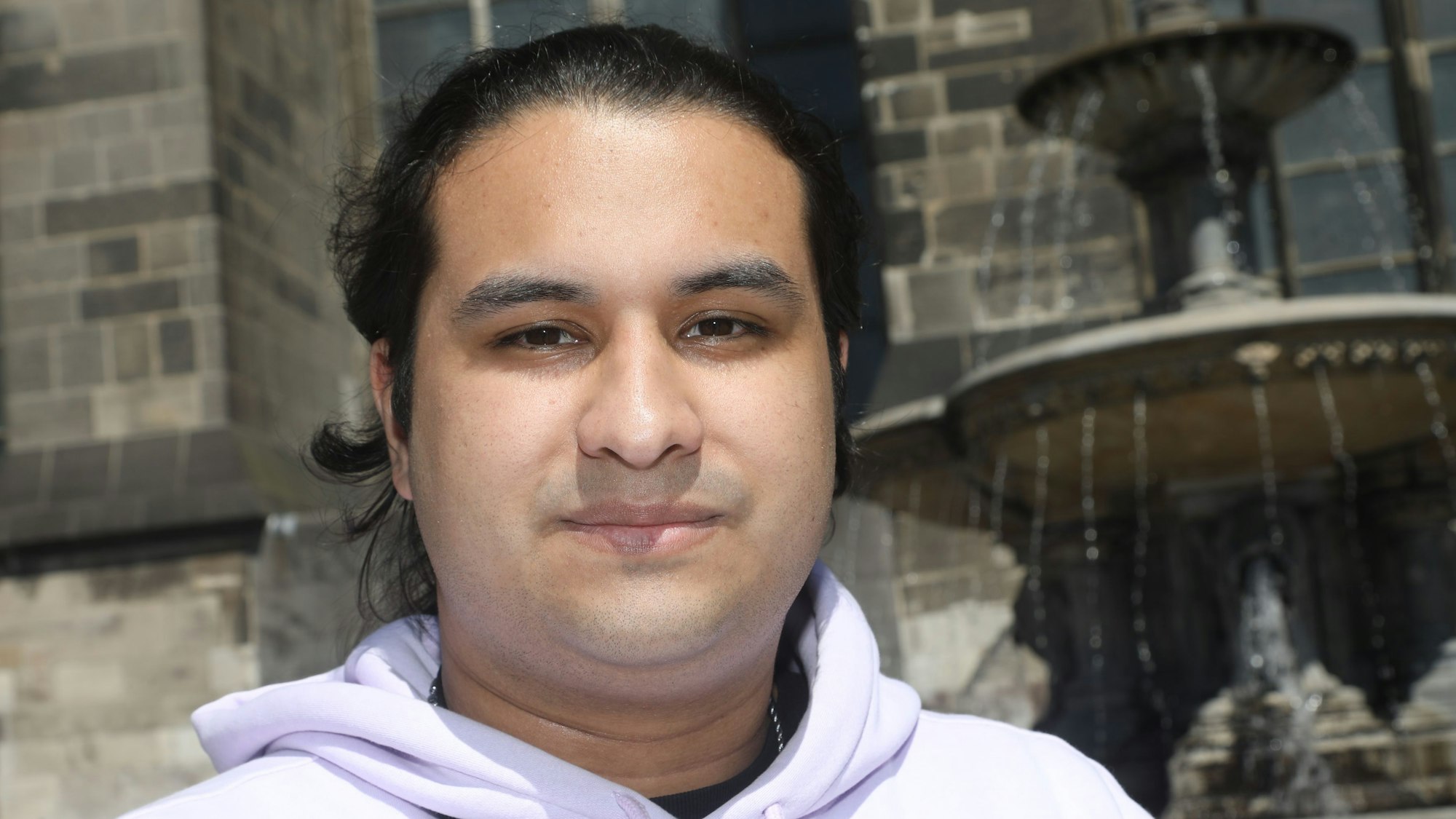 Juan José steht vor dem Kölner Dom und schaut in die Kamera. Rechts hinter ihm sprudelt ein Brunnen.