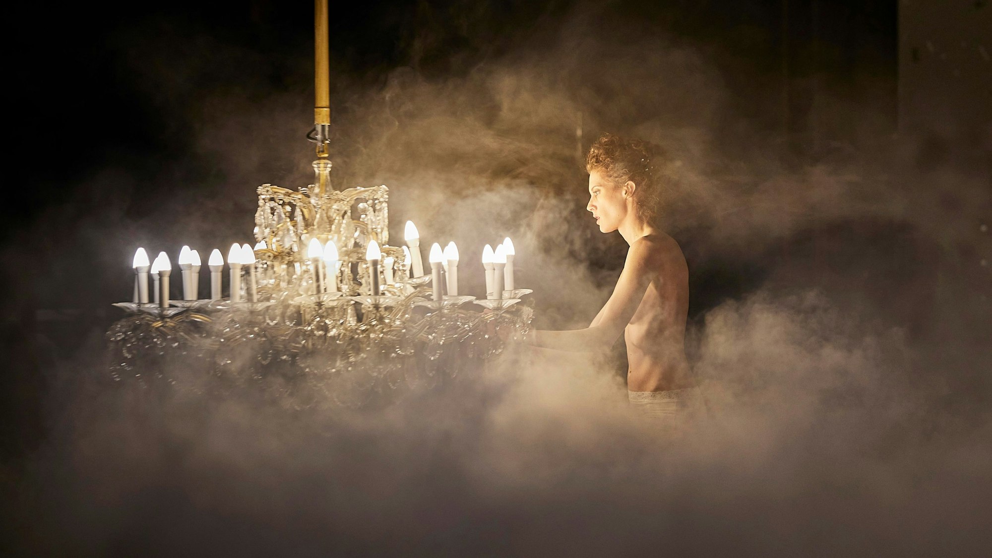 Julia Riedler steht mit bloßem Oberkörper neben einem Kronleuchter im Nebel