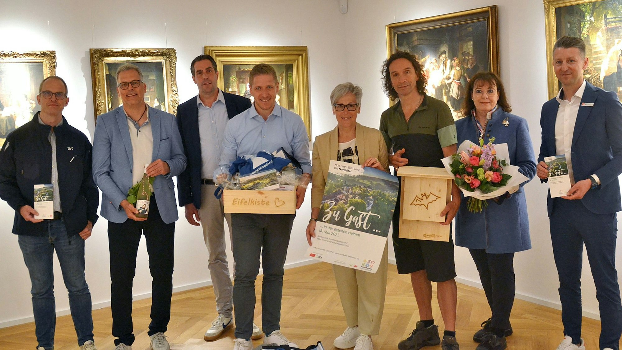 Sechs Männer und zwei Frauen stehen vor einer Reihe Gemälde und halten Blumen, Plakate und Flyer in die Kamera.