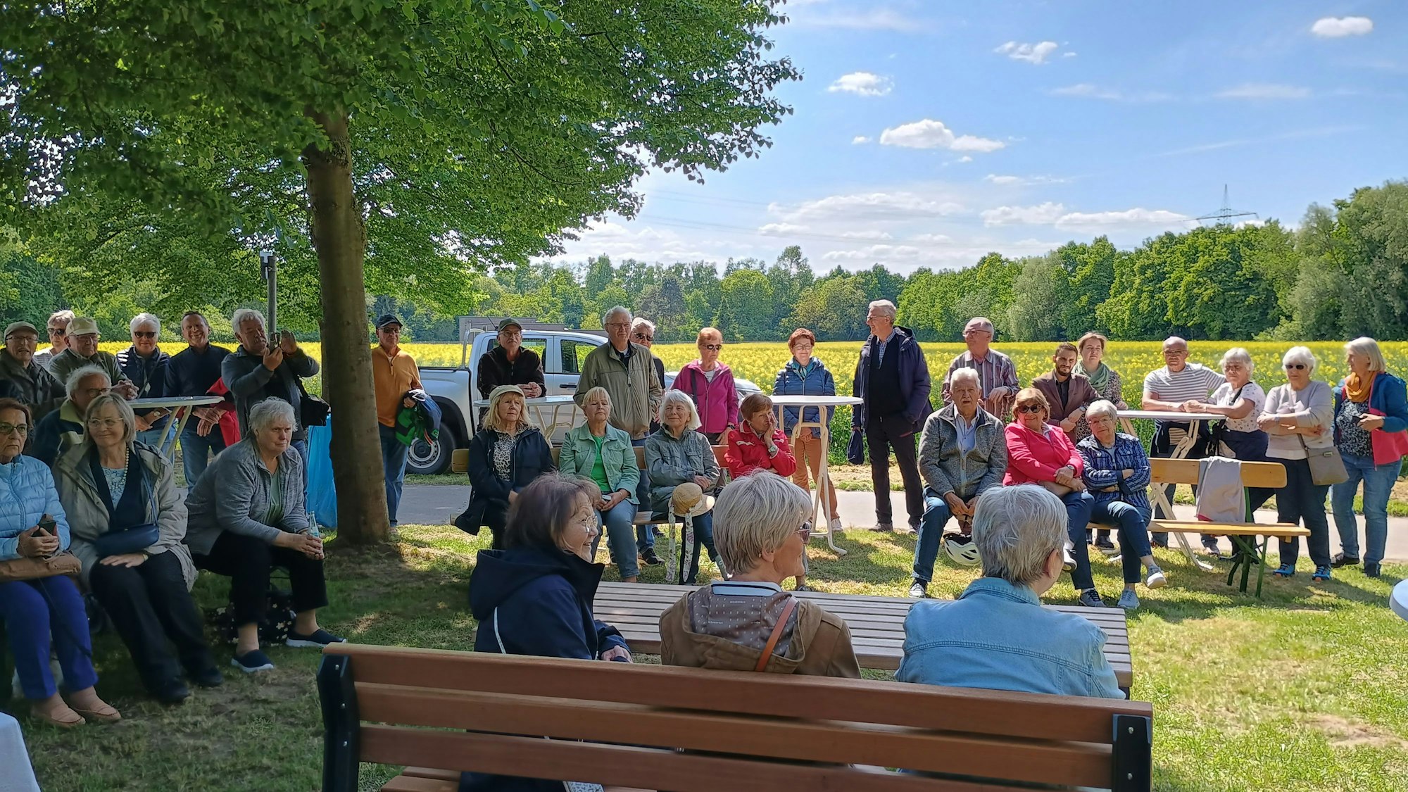 Zu sehen sind die Gäste, die um das Lothringer Kreuz herum auf Bänken Platz genommen haben oder stehen. Im Hintergrund ein gelb blühendes Feld.