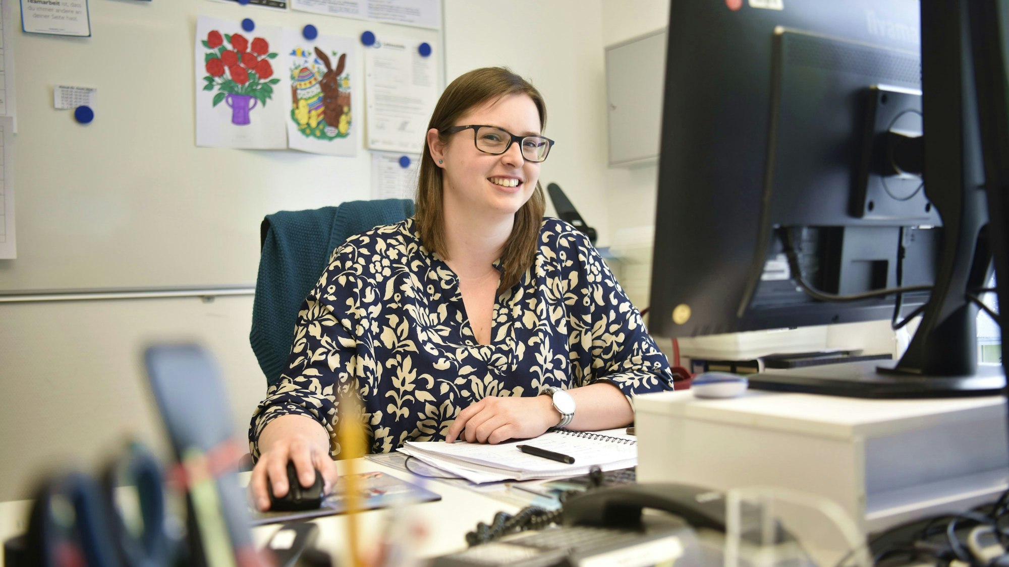 Eine Frau in einem geblümten Kleid sitzt an einem Schreibtisch und arbeitet an einem Bildschirm, im Hintergrund hängen Bilder und Zettel.