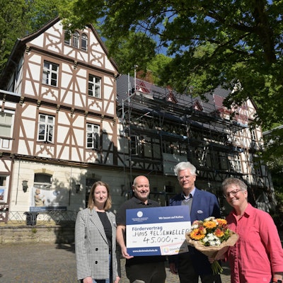 Vier Menschen stehen mit einem großen Scheck vor einem historischen Fachwerkgebäude.